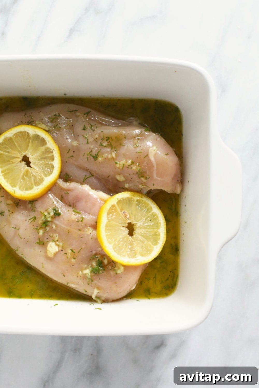 Raw chicken breasts soaking in a vibrant lemon dill marinade in a glass baking dish, ready for cooking.