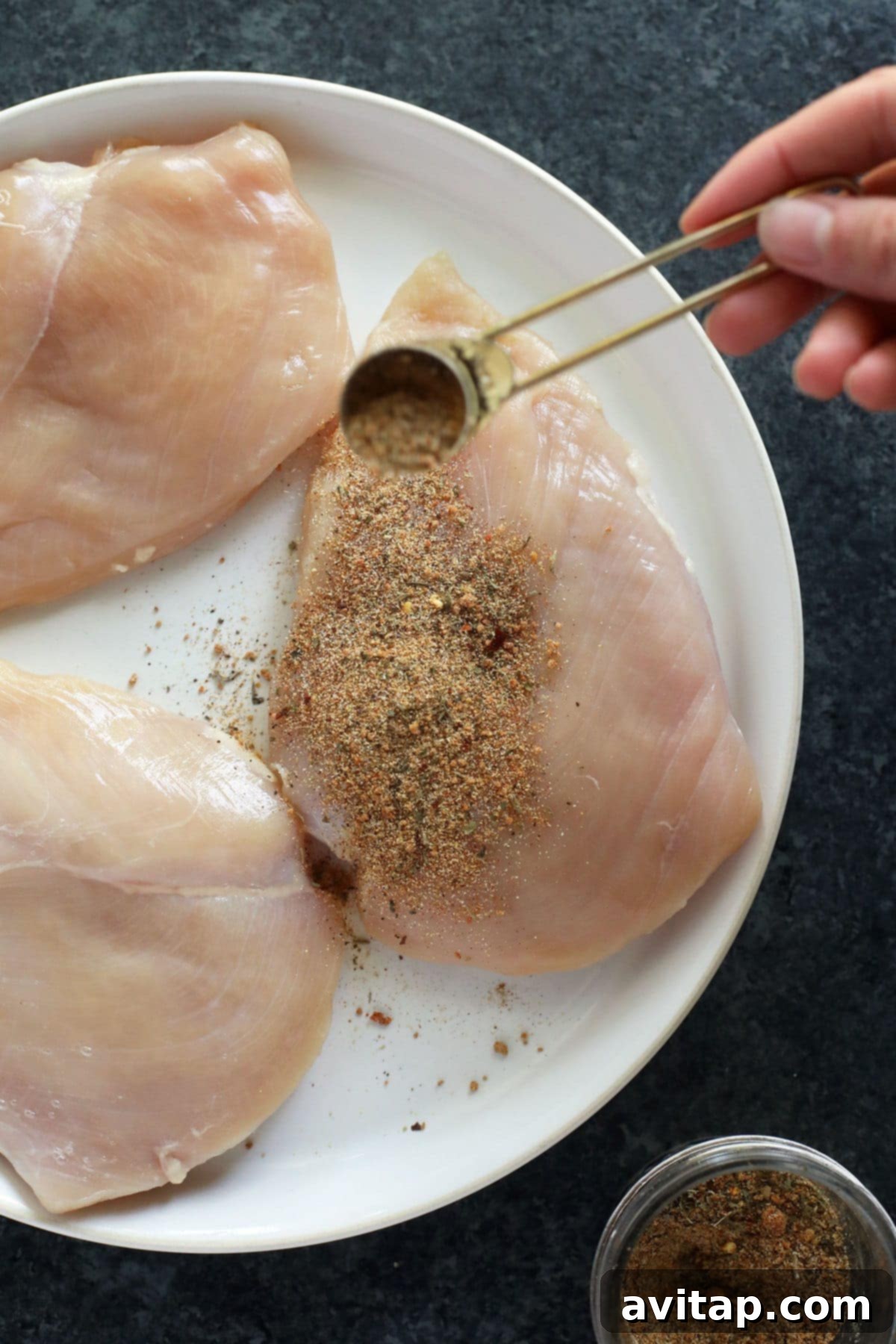 A hand is shown sprinkling a generous amount of all-purpose seasoning onto a raw chicken breast, ensuring full coverage for maximum flavor.