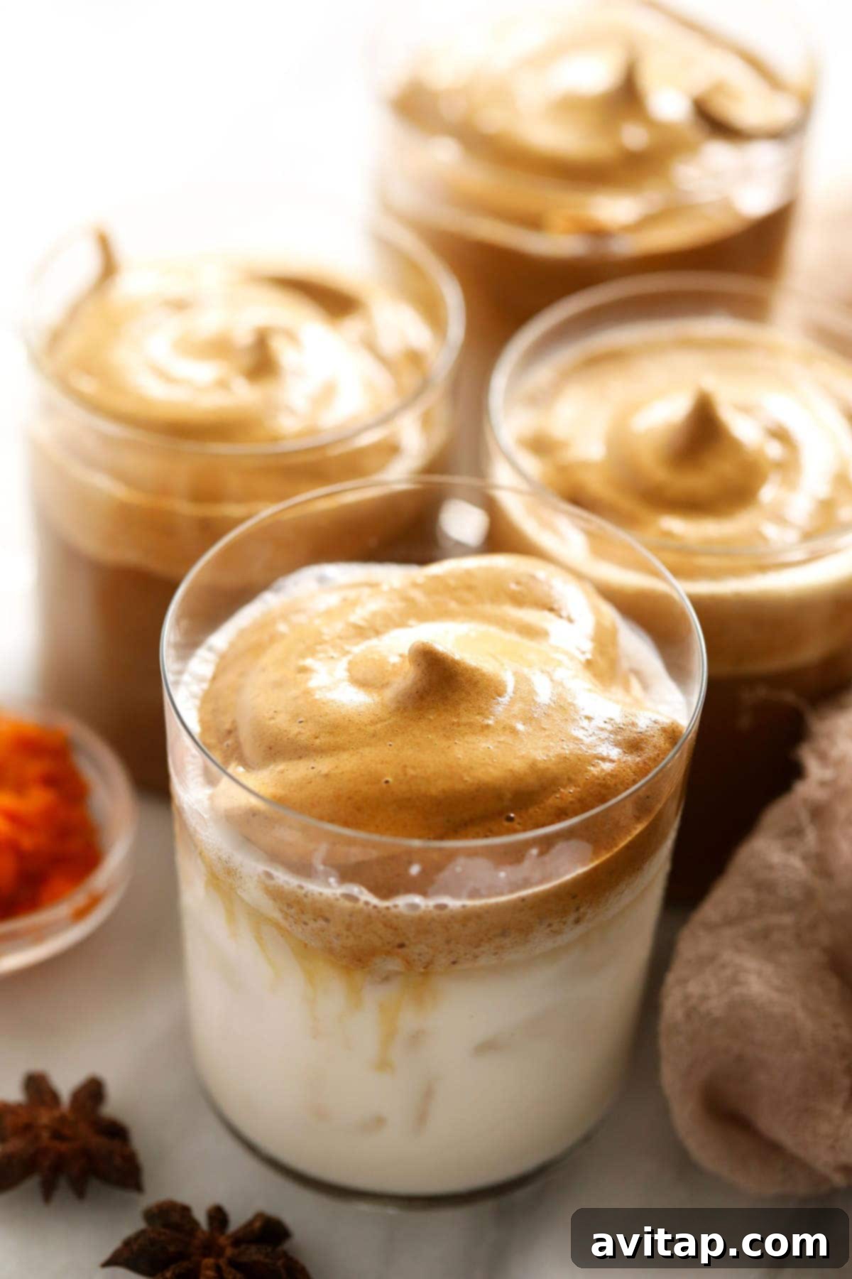 Four cups of pumpkin spice dalgona coffee, garnished with cinnamon sticks and a sprinkle of spice, set on a rustic wooden surface, highlighting the creamy whipped topping and vibrant orange hue.