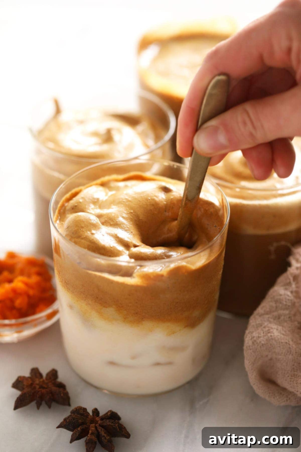 A spoon swirling whipped pumpkin spice dalgona coffee into a glass of almond milk, creating an inviting marble effect.