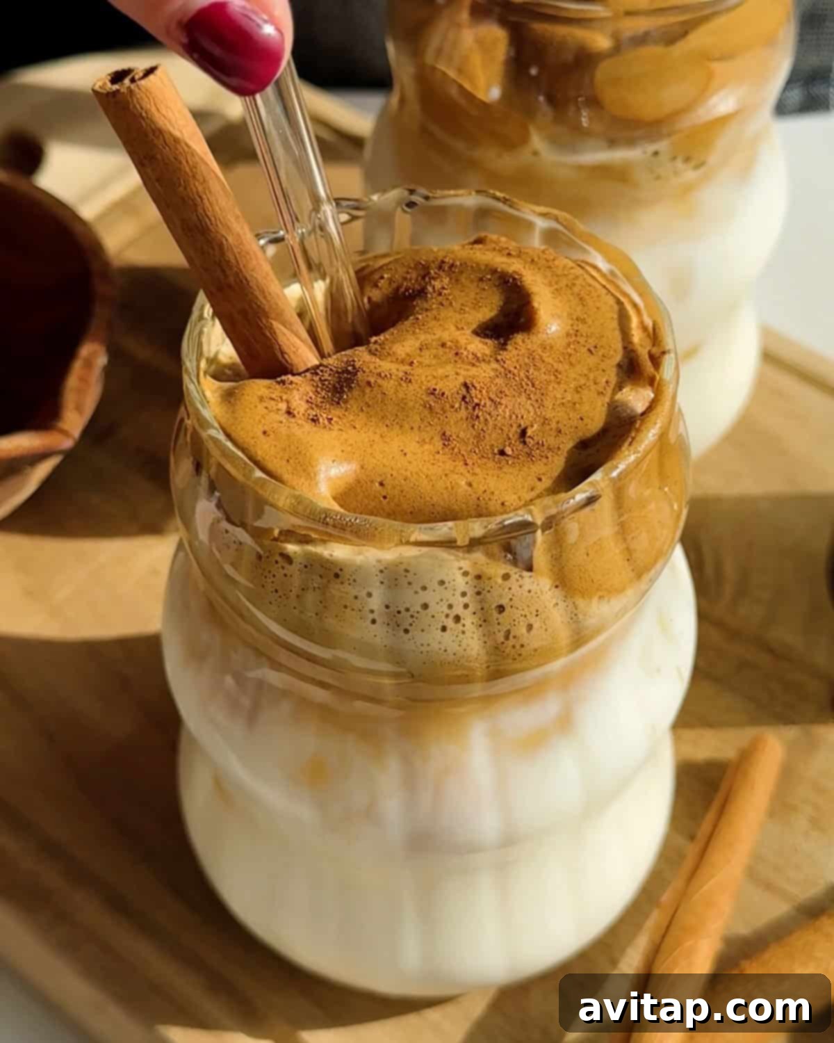 A glass of whipped coffee with a thick foam layer on top, garnished with a cinnamon stick, sits on a wooden board next to a second similar drink.