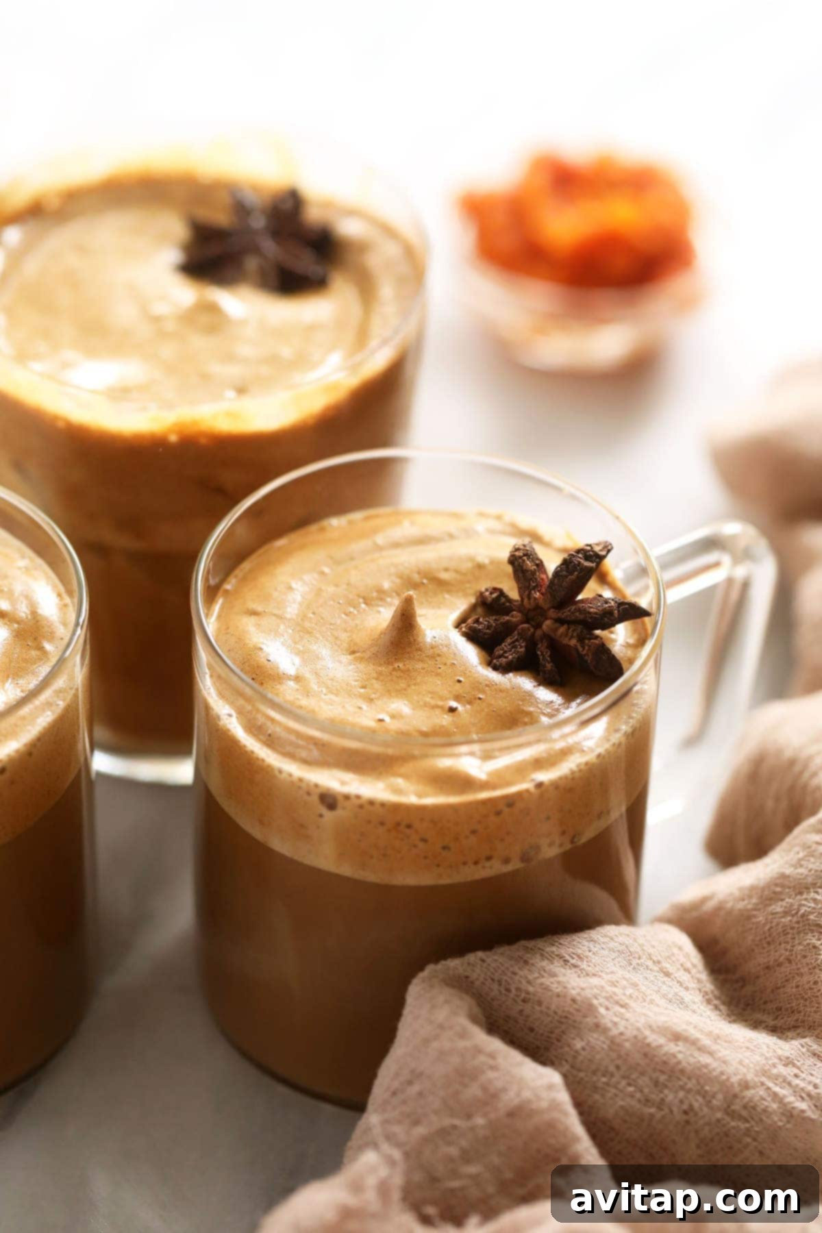 Two mugs of hot dalgona coffee, topped with creamy foam and a sprinkle of pumpkin spice, placed on a cozy knitted mat beside a small pumpkin.