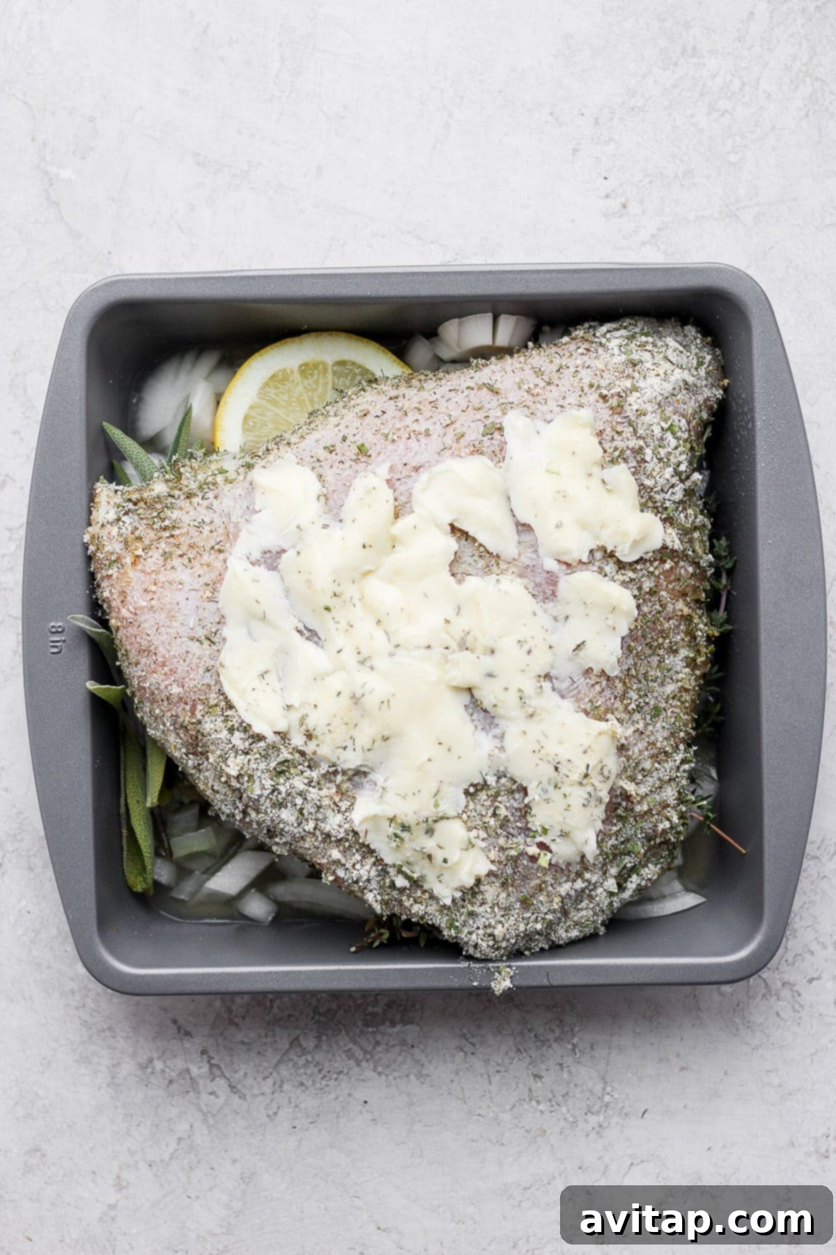 A turkey breast in a baking dish surrounded by butter, lemons, and fresh herbs.