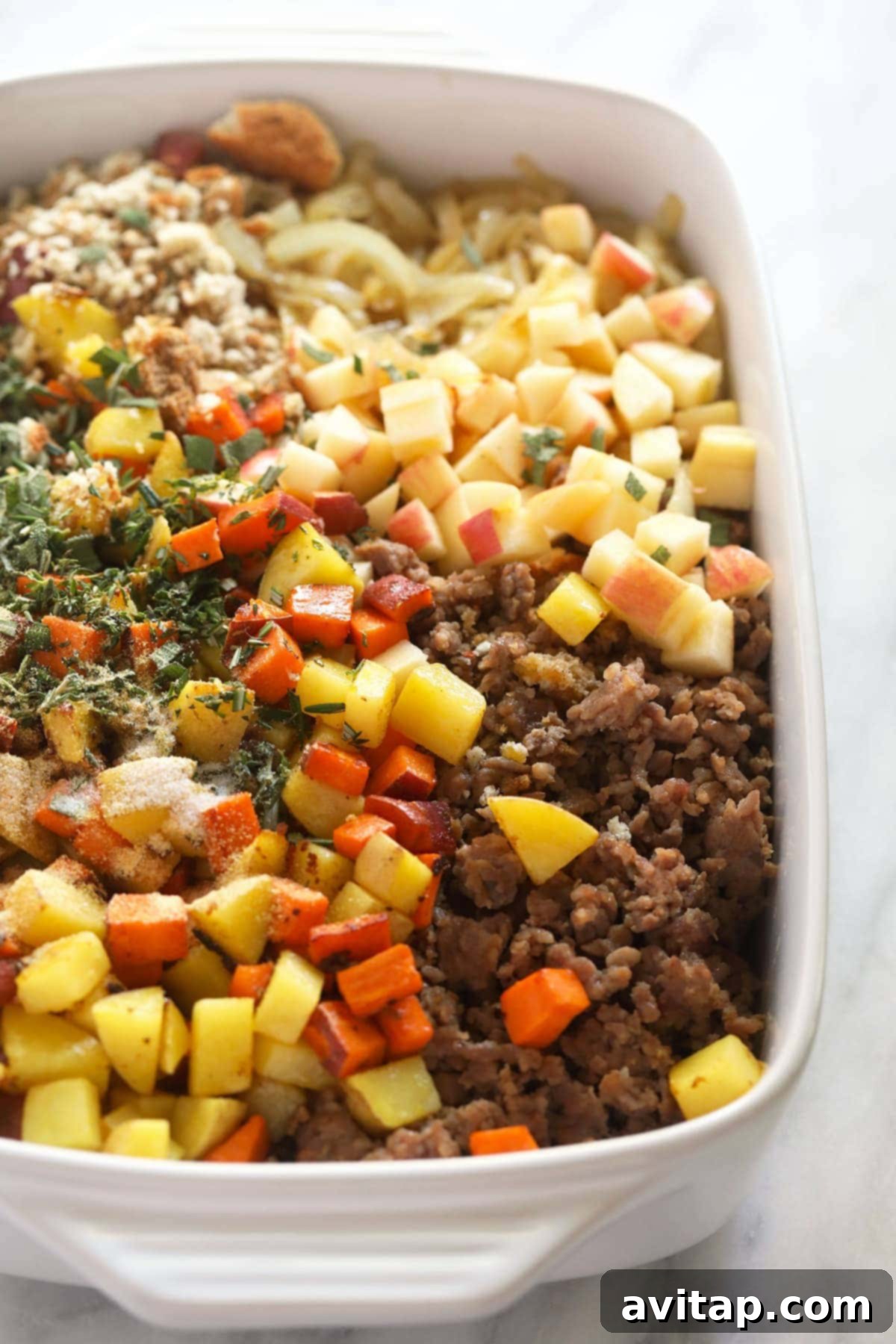 All the fresh ingredients for sausage stuffing, including Italian sausage, potatoes, apples, onions, and fresh herbs, arranged in a casserole dish before mixing.