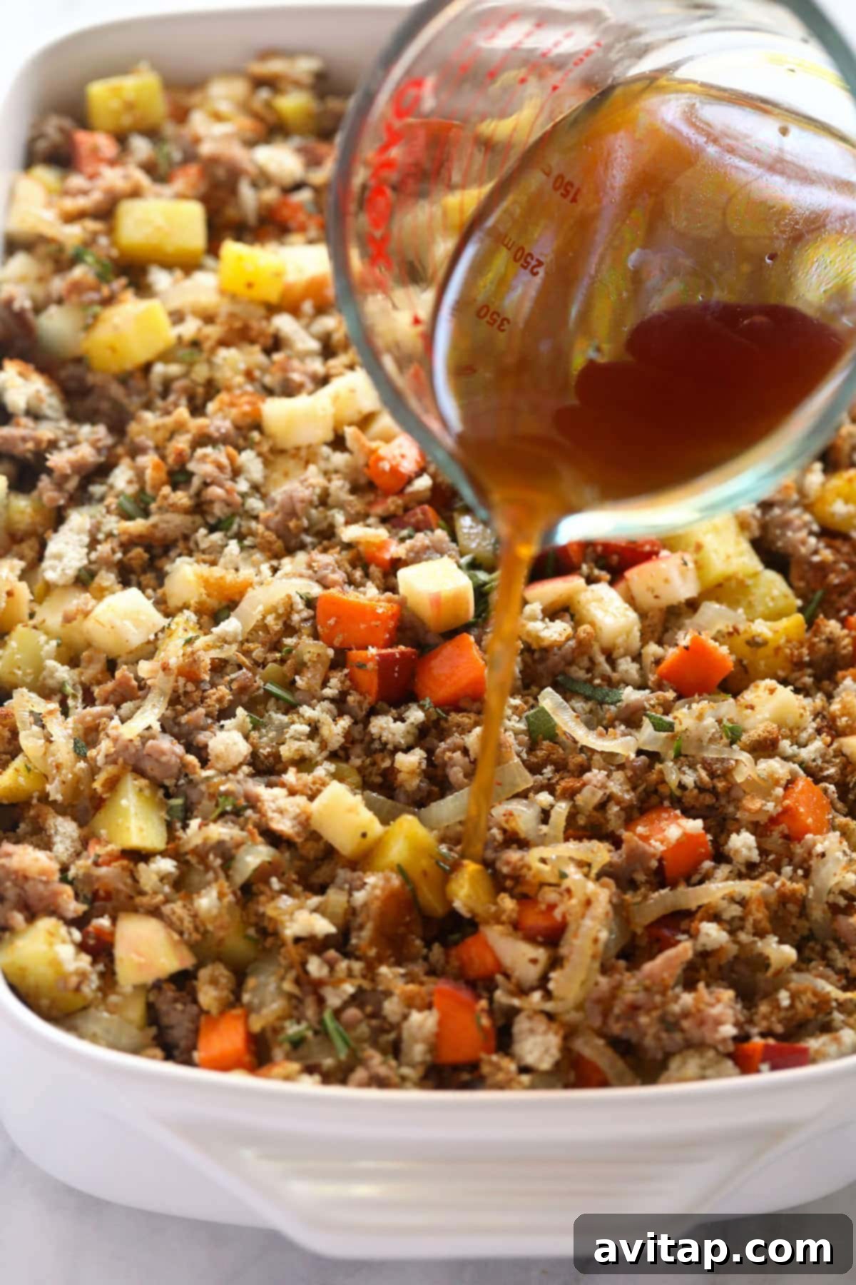 Rich beef broth being poured evenly over the mixed sausage stuffing in a casserole dish before baking.