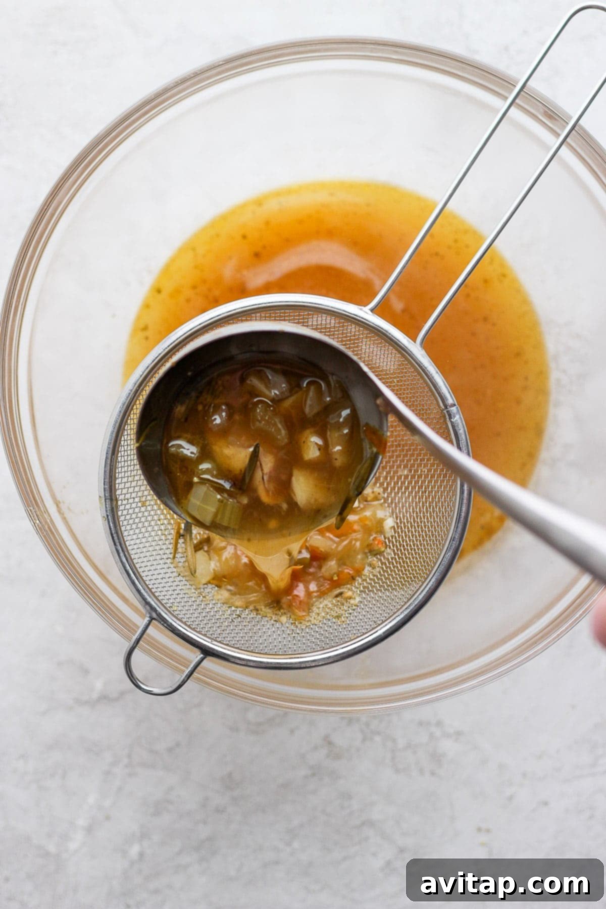 A person carefully pouring strained chicken broth through a fine-mesh sieve into a clean bowl.