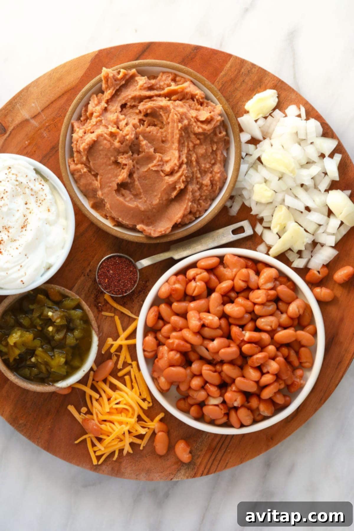 Various fresh ingredients for homemade bean dip, including cans of beans, a block of cheddar cheese, Greek yogurt, salsa, and minced aromatics, arranged neatly on a wooden board before mixing.
