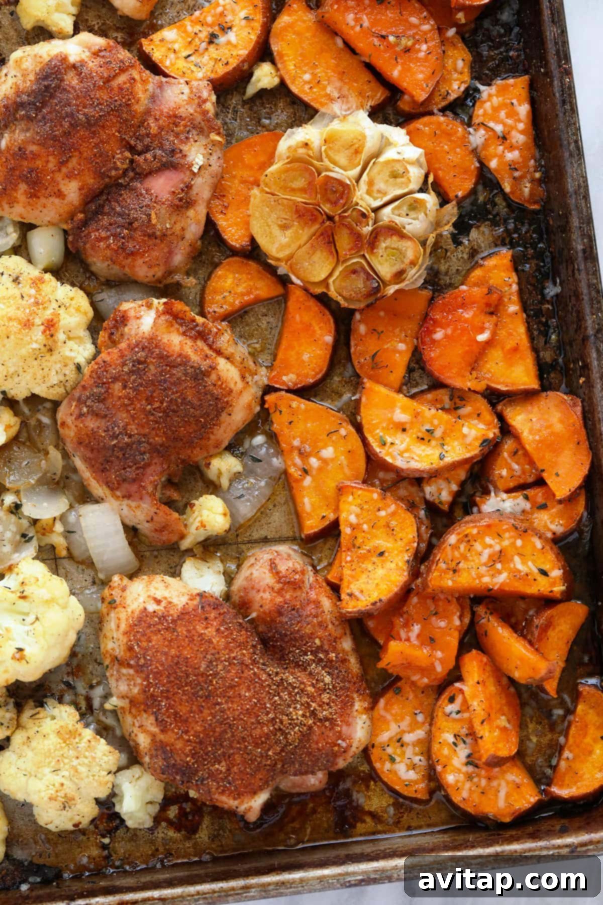 Golden brown chicken, sweet potatoes, cauliflower, onion, and a roasted head of garlic artfully arranged on a baking sheet, ready to be served.