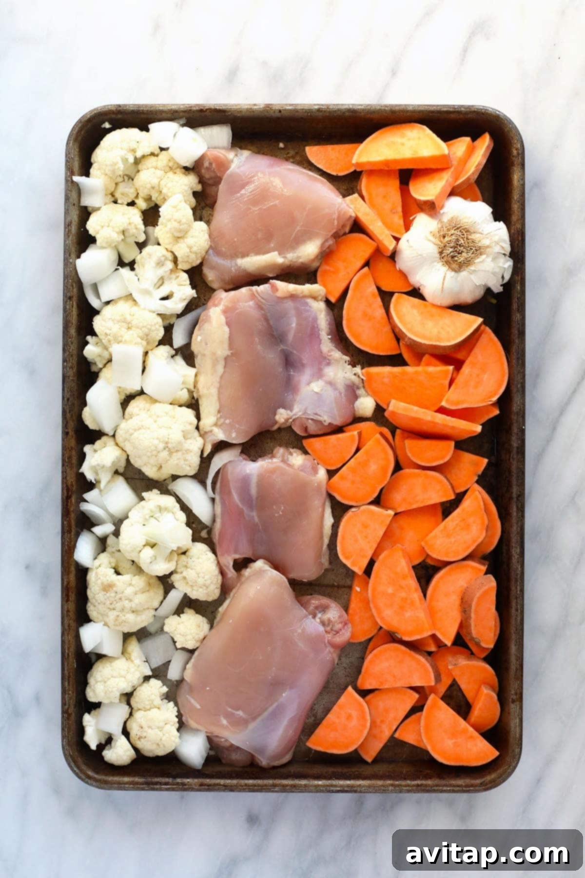 Raw chicken thighs, vibrant orange sweet potato chunks, white cauliflower florets, sliced red onion, and a head of garlic neatly arranged on a baking sheet, prepped and ready for roasting.