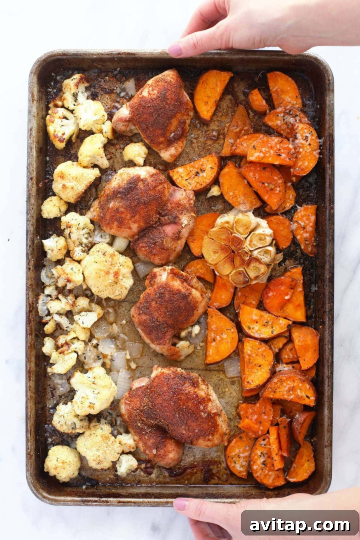 Close-up of baked chicken thighs with crispy skin, perfectly roasted garlic, tender cauliflower, and caramelized sweet potatoes on a baking sheet, fresh out of the oven.