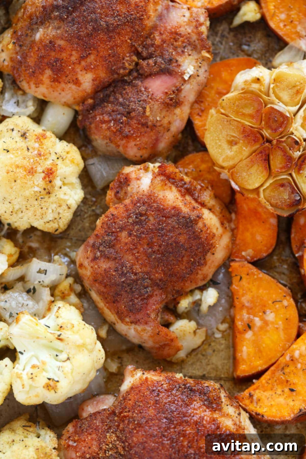 A close-up shot of a perfectly baked sheet pan meal, featuring succulent chicken thighs, golden-brown roasted garlic, tender cauliflower, and caramelized sweet potatoes, ready to be divided into portions.