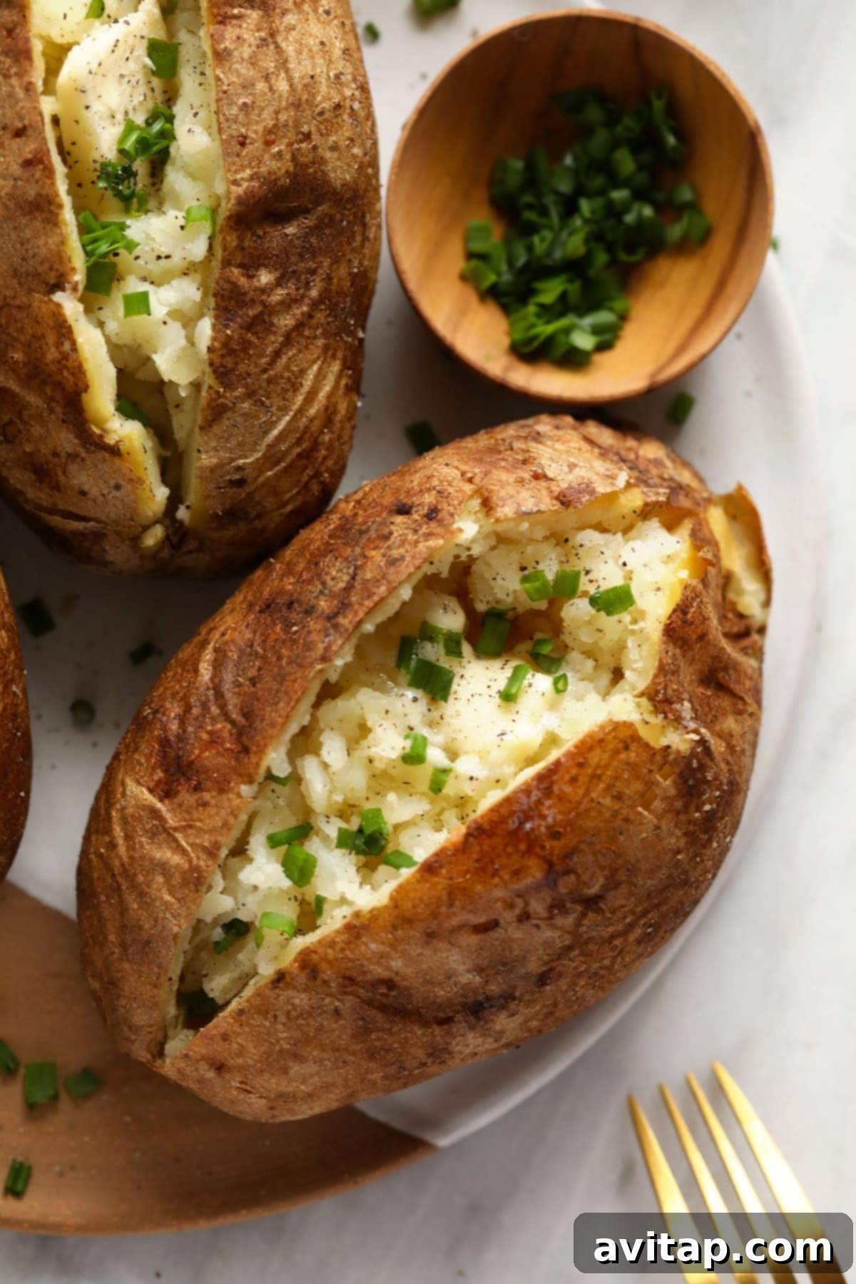 The Ultimate Crispy Fluffy Baked Potato 2 Baked potatoes on a plate with butter, salt, pepper, and chives, showcasing their crispy skin and fluffy texture.
