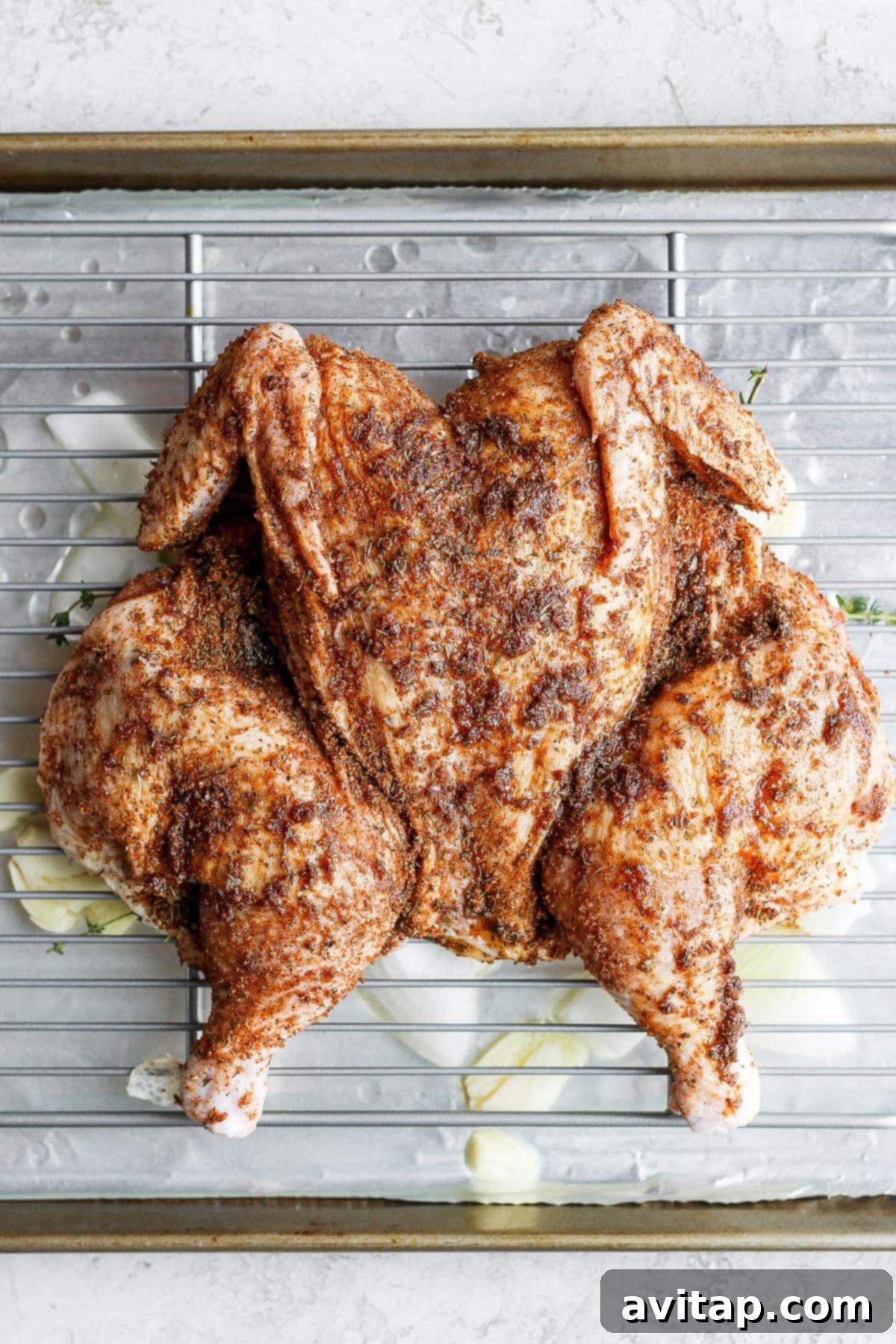 A raw spatchcock chicken perfectly flattened and placed on a wire rack within a baking sheet, seasoned and ready for the oven.