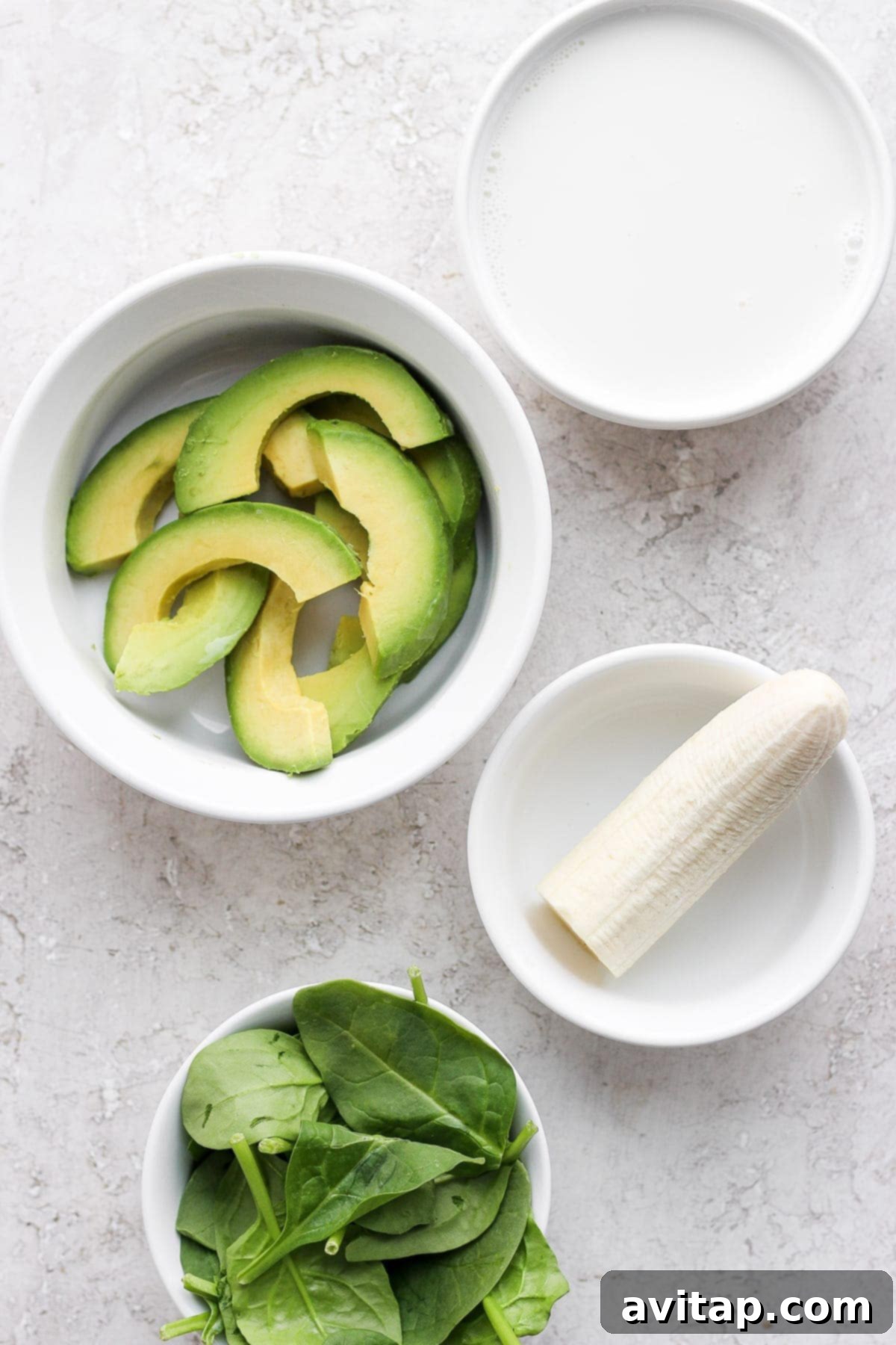 Essential Avocado Smoothie Ingredients Individual ingredients for an avocado smoothie laid out in bowls, including frozen avocado, spinach, banana, and almond milk, ready to be added to a blender.