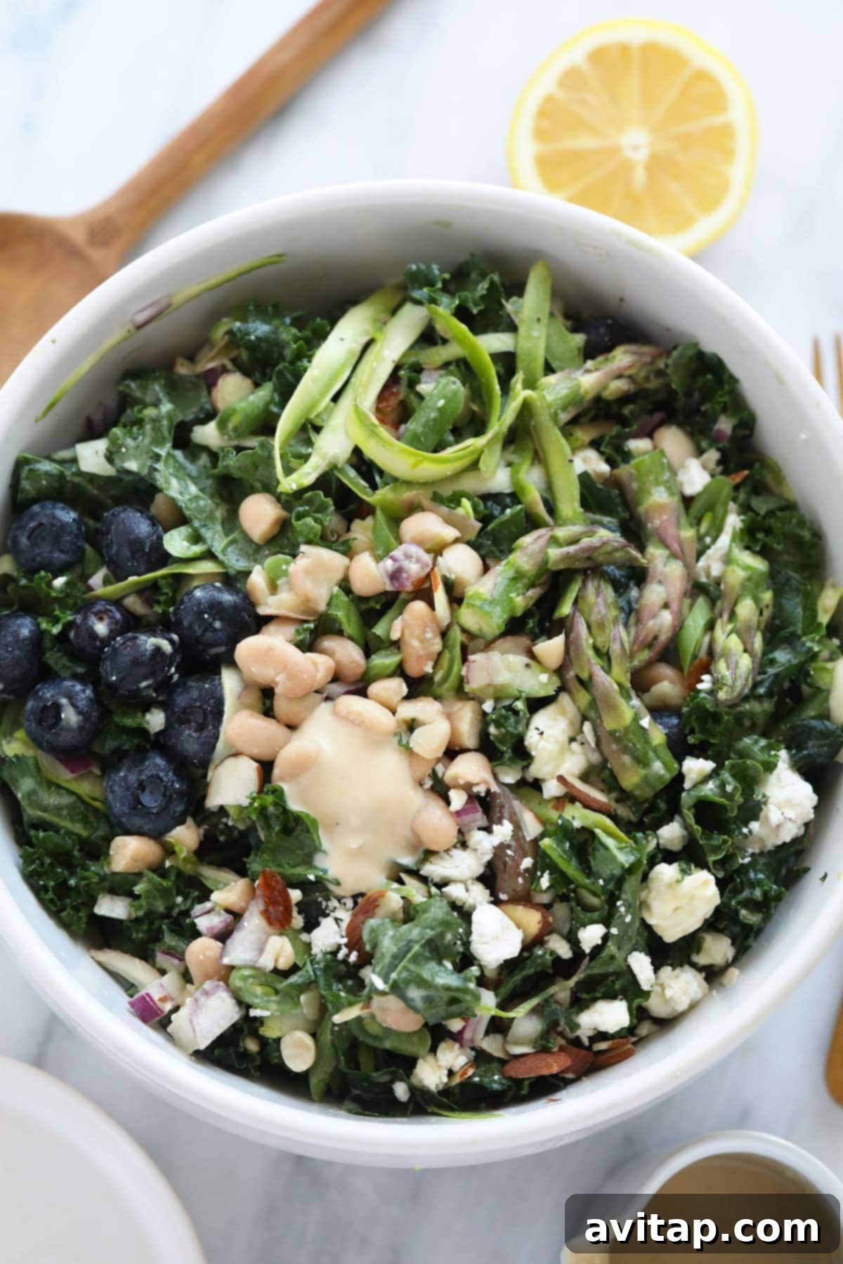 A large, vibrant bowl of asparagus salad, brimming with shaved asparagus, kale, blueberries, beans, and feta, artfully placed on a wooden table, ready to be served at a gathering.