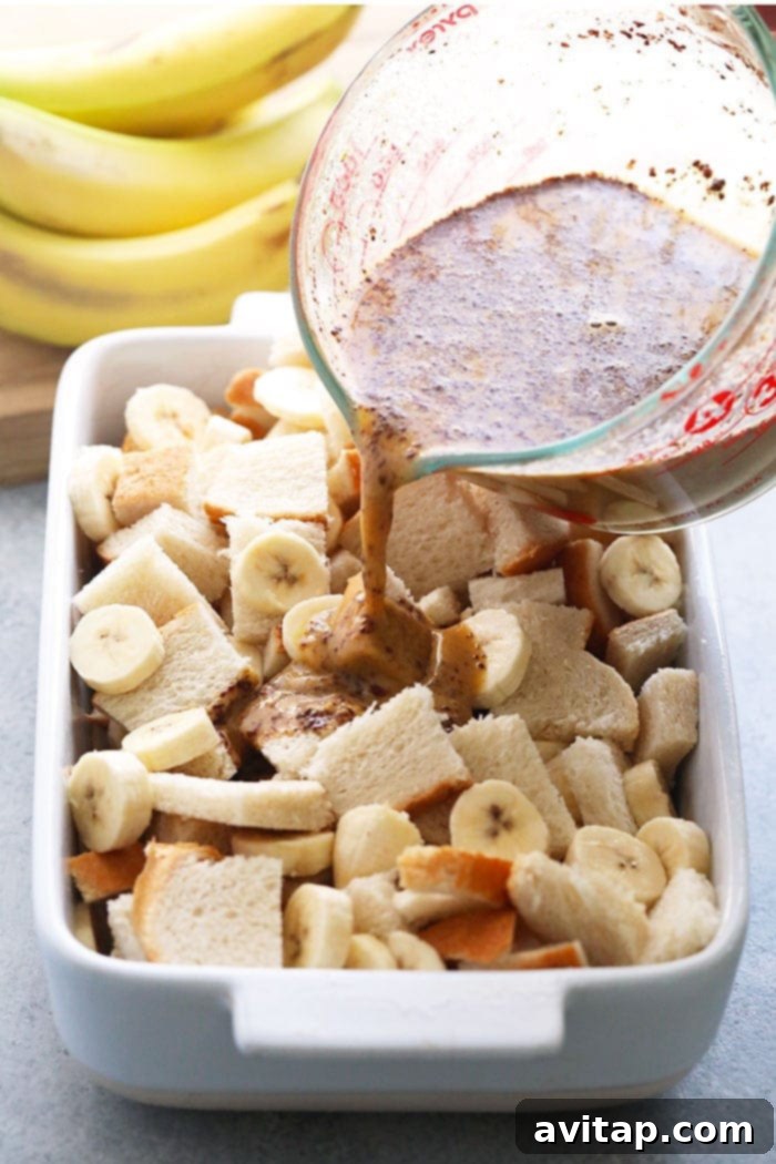 Hands pouring peanut butter banana custard mixture over cubed bread and sliced bananas in a casserole dish.