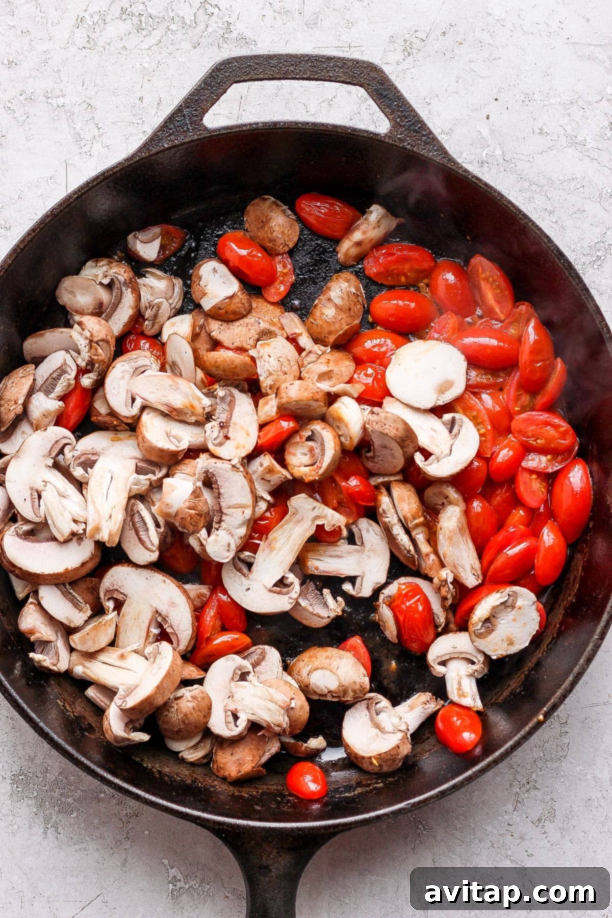Sliced Bella mushrooms and halved cherry tomatoes being sautéed in a cast iron skillet, glistening with olive oil.