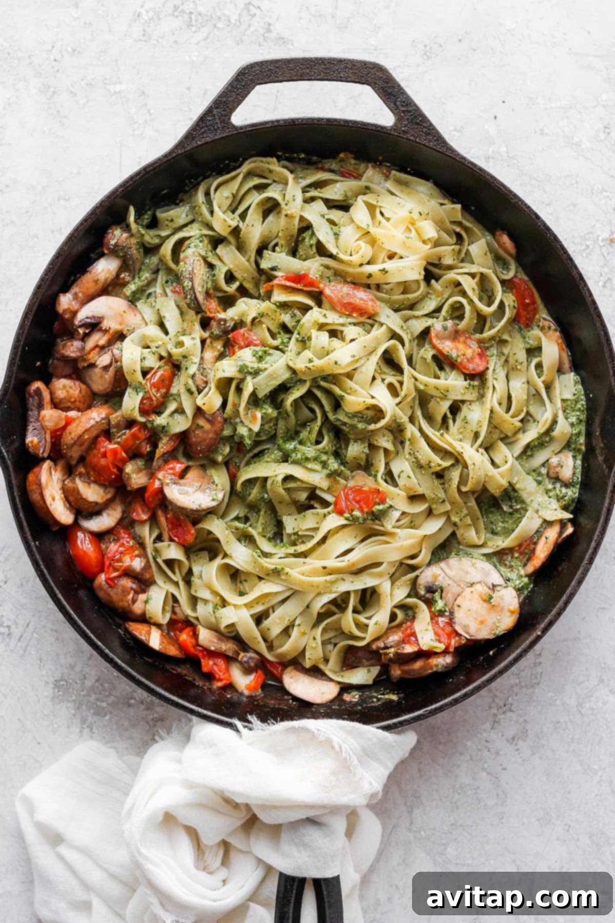 A vibrant bowl of vegan pesto pasta in a rustic cast iron skillet, showcasing fresh basil leaves, cherry tomatoes, and sautéed mushrooms.