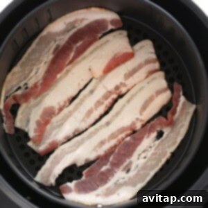 Raw bacon slices being carefully placed into an air fryer basket, ensuring a single layer.