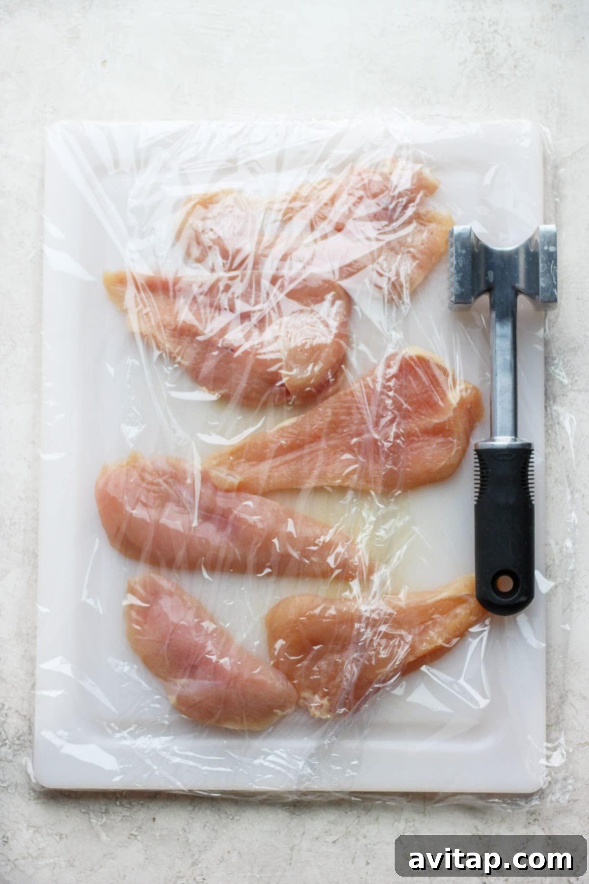 Raw chicken breasts being pounded to uniform thickness on a cutting board, covered with plastic wrap
