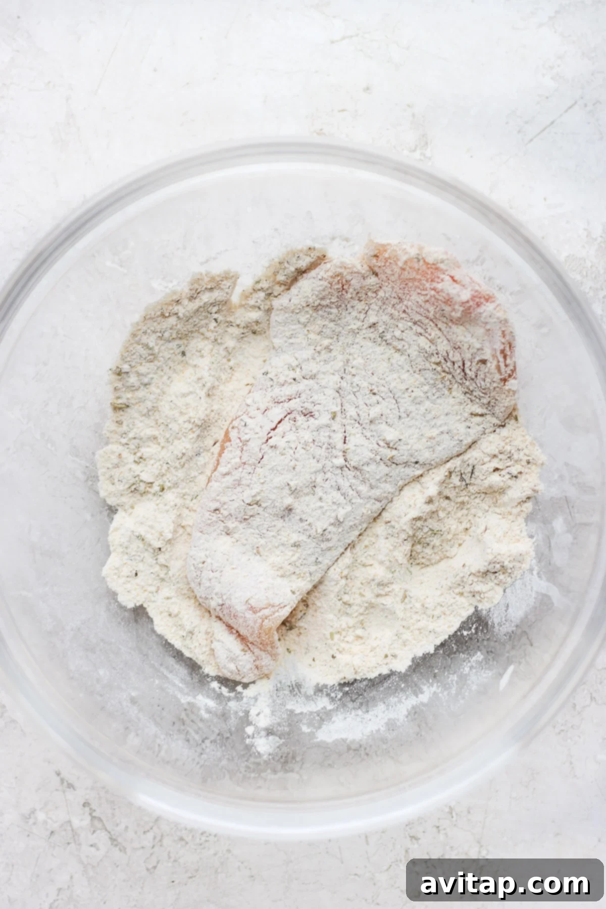 Raw chicken breast being thoroughly dredged in a bowl of seasoned flour mixture