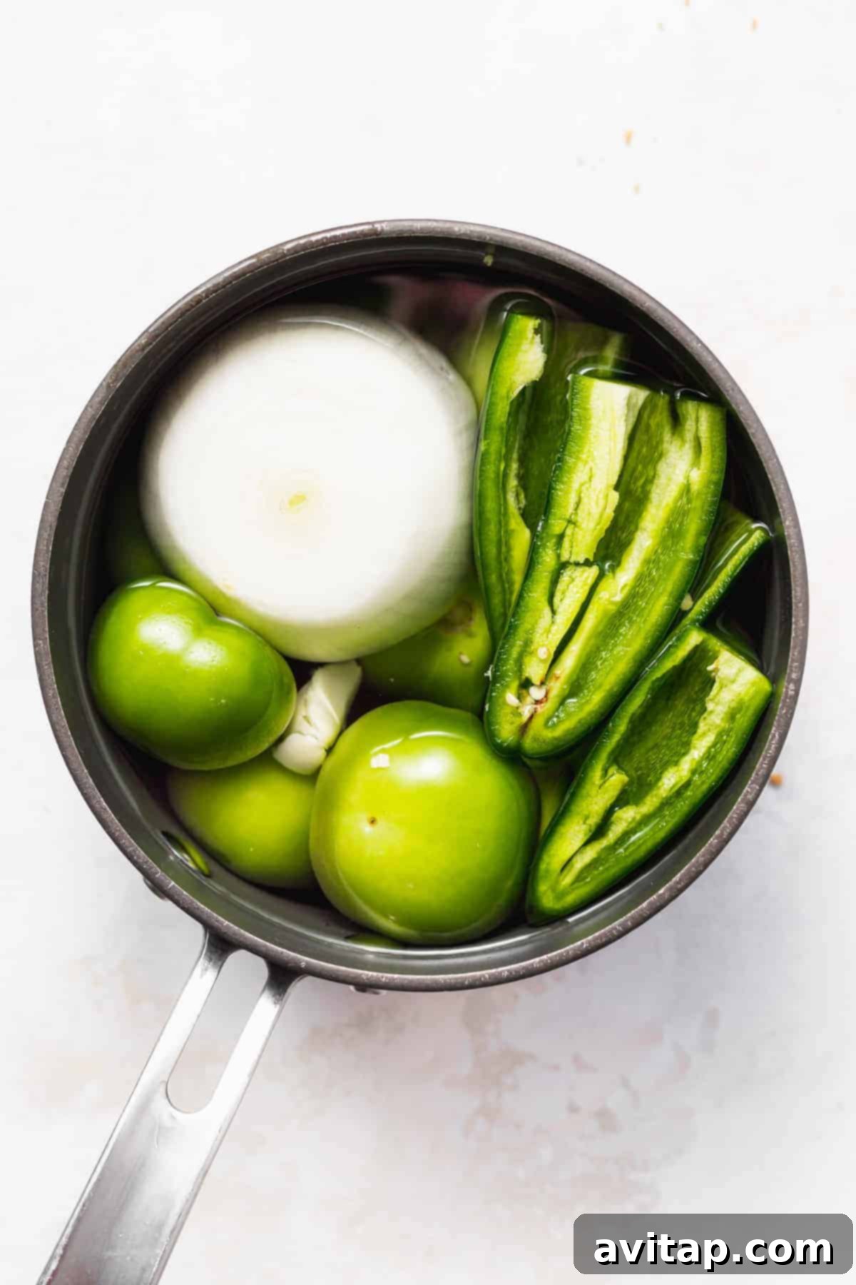 Fresh tomatillos, garlic cloves, white onion, and jalapeños simmering gently in a saucepan with water, forming the flavorful base of the salsa verde.