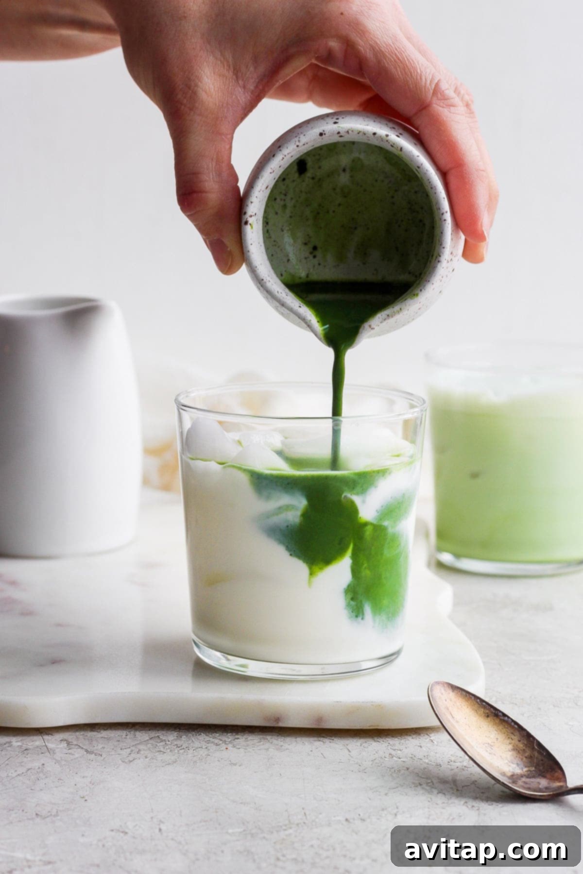 Pouring bright green matcha mixture into a glass of almond milk over ice, creating a beautiful layered iced matcha latte.
