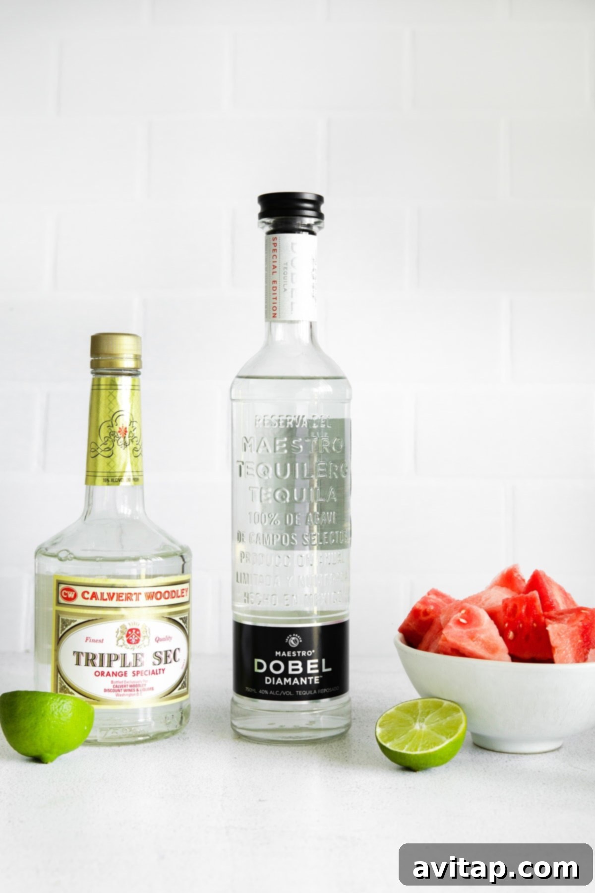 Essential watermelon margarita ingredients including Triple Sec, tequila, fresh watermelon chunks, and lime wedges artfully arranged on a kitchen countertop.