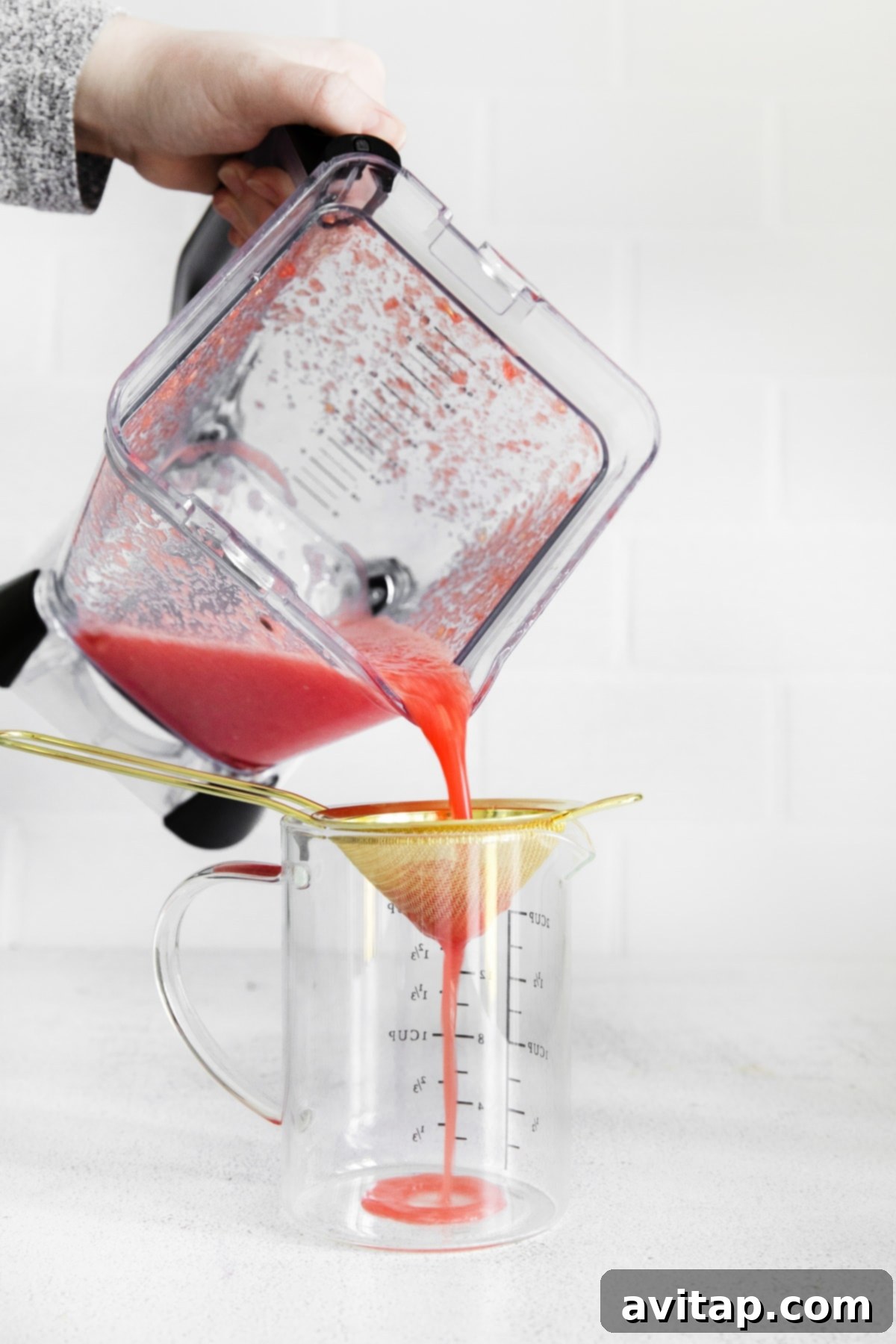 An action shot of vibrant blended watermelon being poured through a fine-mesh sieve to extract smooth watermelon juice, a key step for a refined margarita.