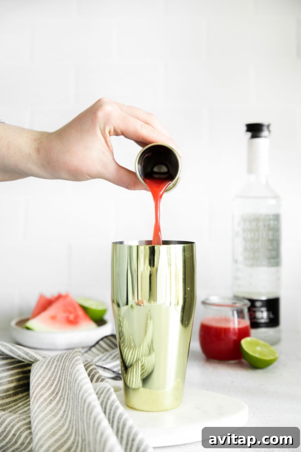 A cocktail shaker on a counter, with a hand pouring freshly made watermelon juice into it, alongside other margarita ingredients.