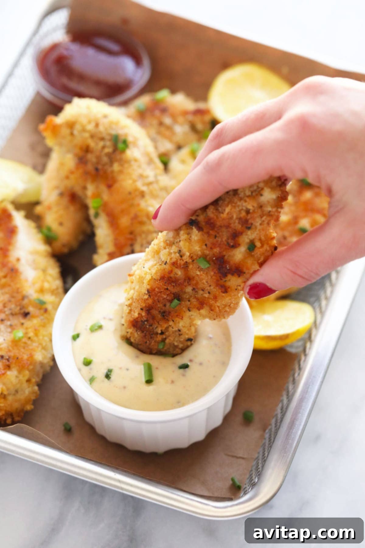Golden Baked Chicken Fingers 7 A hand dipping a crispy baked chicken tender into a bowl of creamy honey mustard sauce.