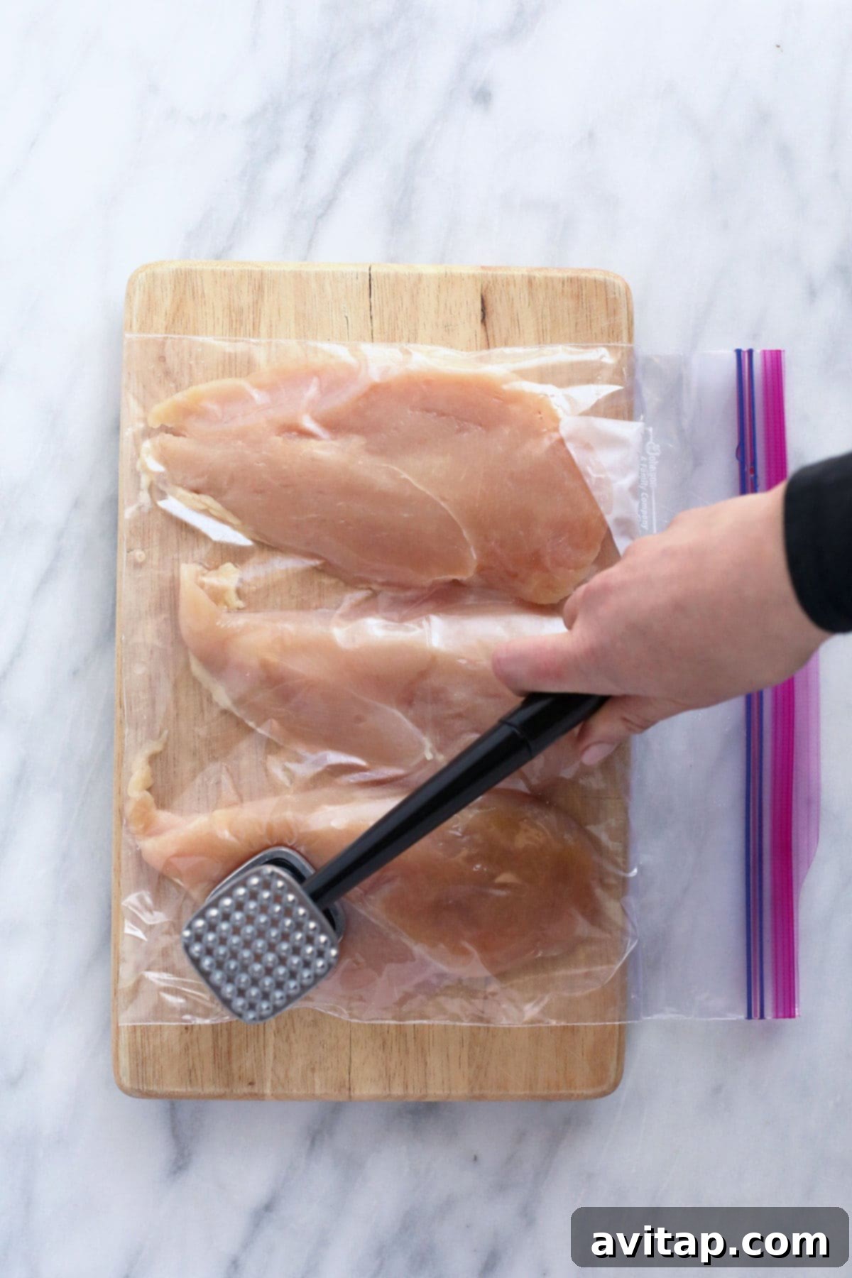 Golden Parmesan Chicken 3 A hand using a meat tenderizer to pound chicken breasts between plastic wrap on a cutting board, ensuring even thickness for cooking.