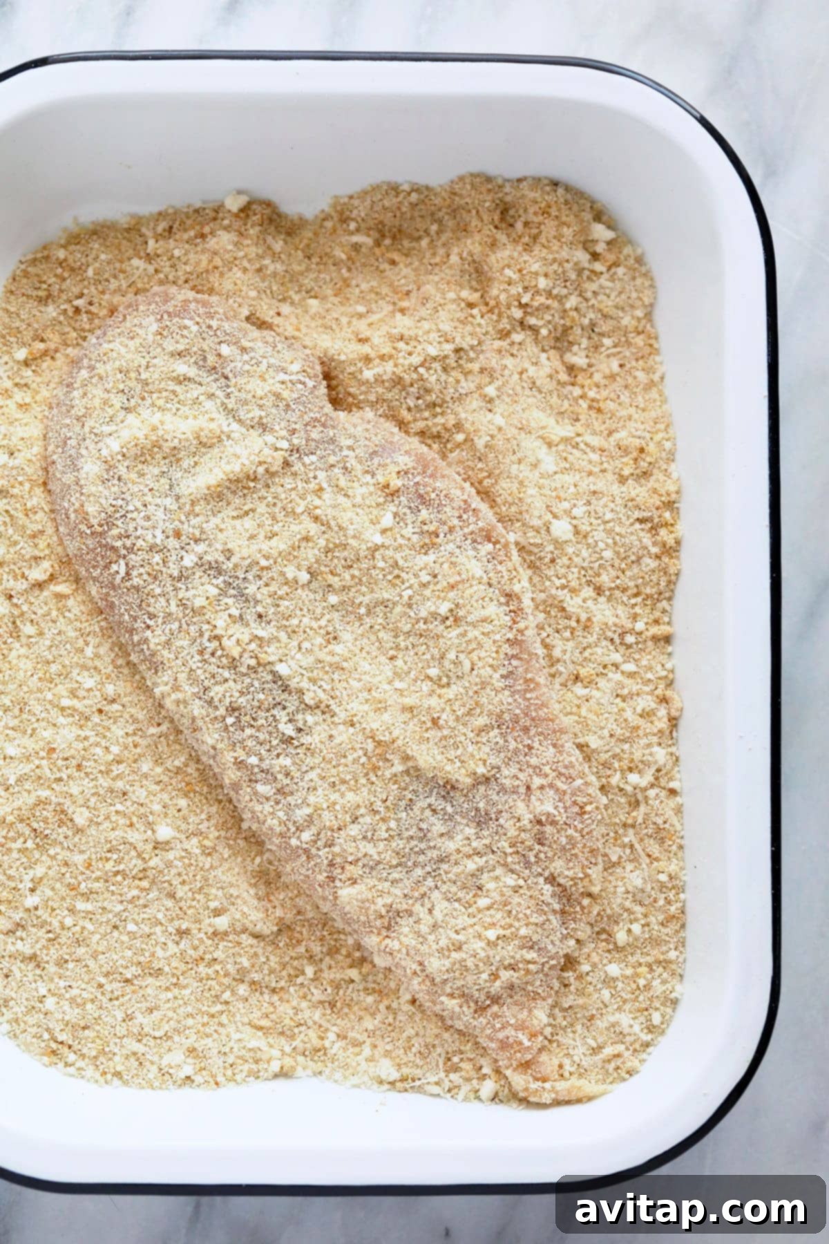 Golden Parmesan Chicken 4 A chicken breast being dredged in a shallow bowl containing a mixture of breadcrumbs and grated parmesan cheese, showing thorough coating.