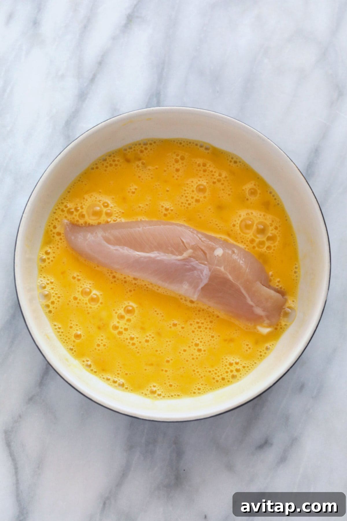 A chicken tenderloin being dipped into a bowl of egg wash, ensuring full coverage