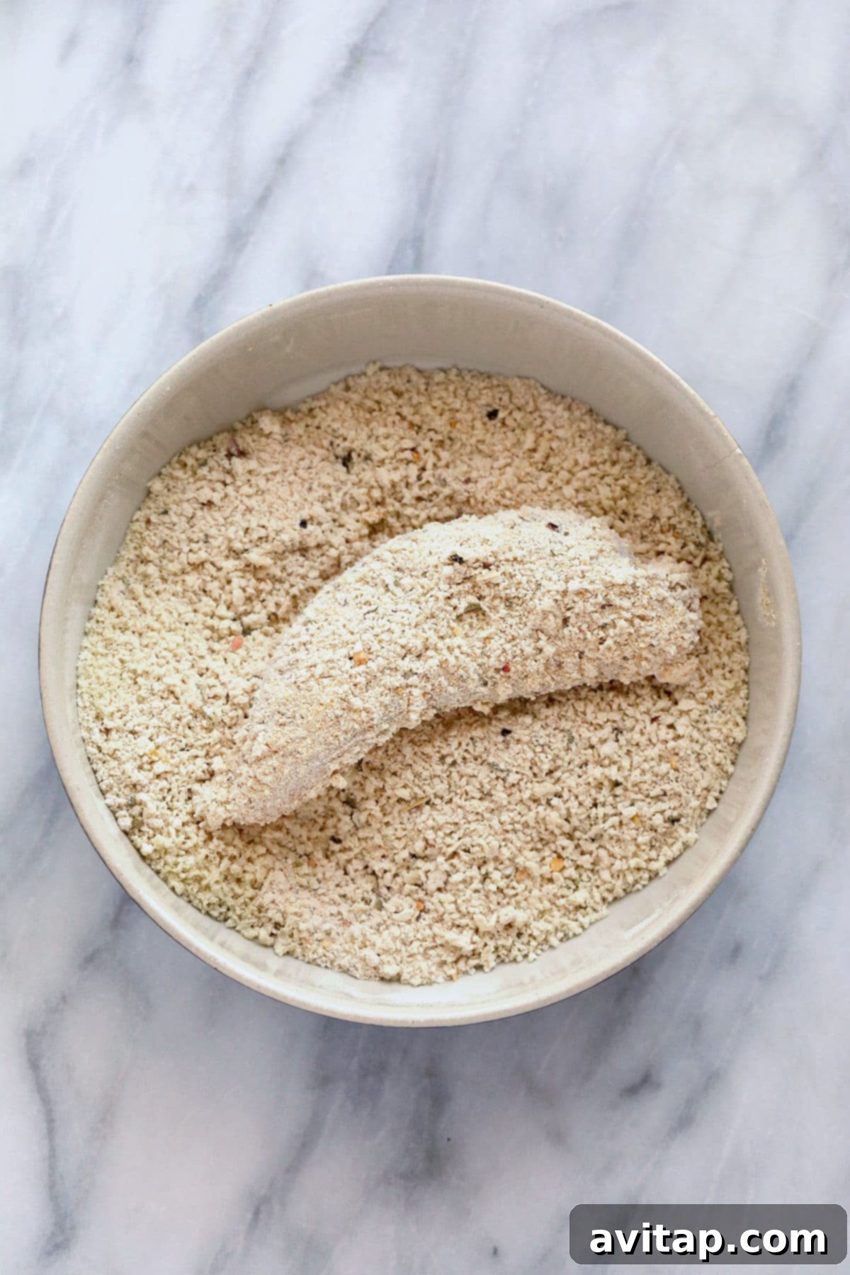 A hand dredging a chicken tenderloin in a bowl of seasoned breadcrumbs