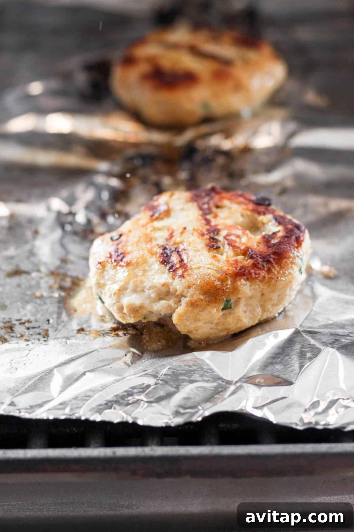 Ground chicken burger patty cooking on tin foil on a grill, showing sizzle and grill marks.