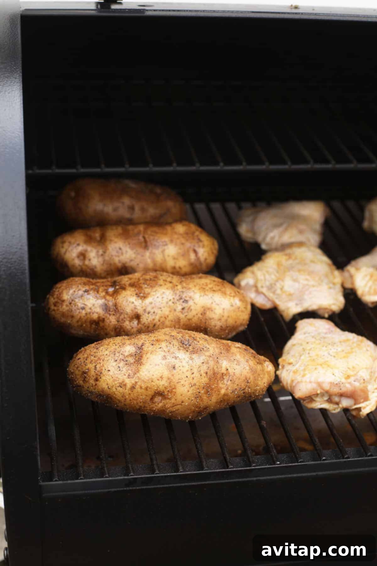 Multiple smoked baked potatoes cooking on a Traeger smoker alongside delicious smoked chicken thighs, showcasing a complete meal being prepared simultaneously.