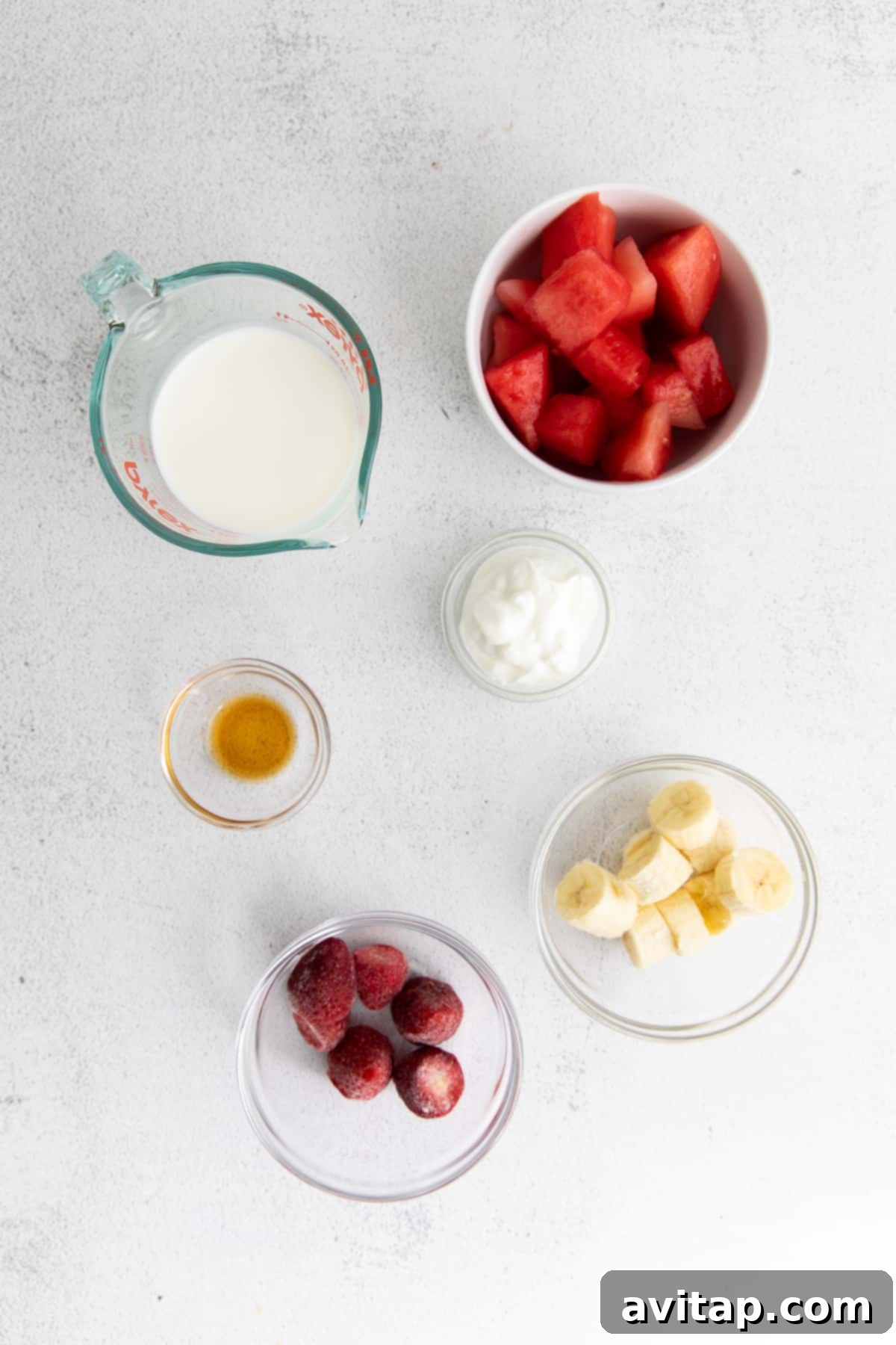 Individual bowls containing fresh watermelon chunks, strawberries, Greek yogurt, banana slices, almond milk, and vanilla extract—all the ingredients needed for a watermelon smoothie.