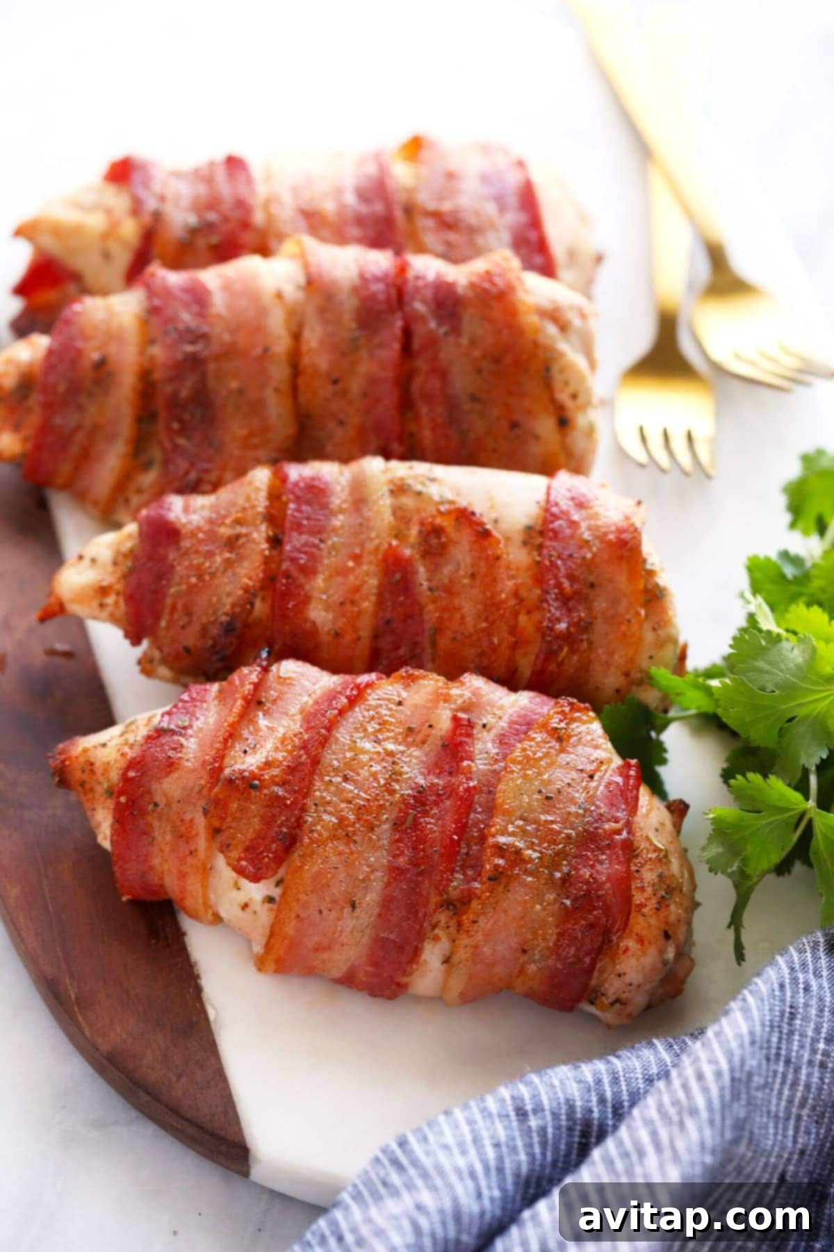 Bacon wrapped chicken is seasoned chicken breasts wrapped in bacon and then baked at high heat for crispy bacon on the outside and tender chicken breast on the inside. Golden brown oven-baked bacon wrapped chicken breasts on a cutting board, garnished with fresh herbs, ready to be served.