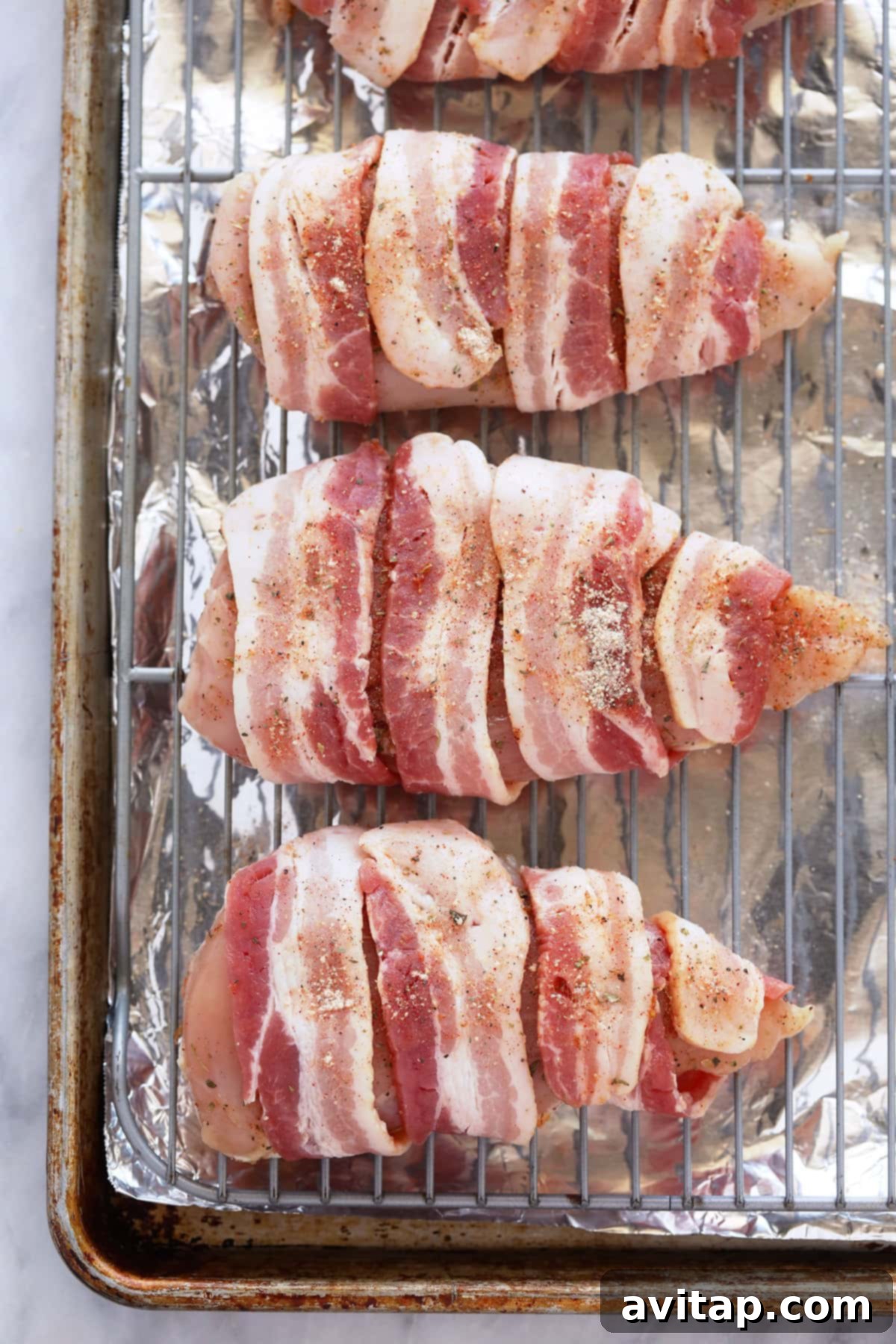 Bacon wrapped chicken is seasoned chicken breasts wrapped in bacon and then baked at high heat for crispy bacon on the outside and tender chicken breast on the inside. Raw, seasoned chicken breasts individually wrapped in strips of uncooked bacon, arranged on a baking sheet, ready for the oven.
