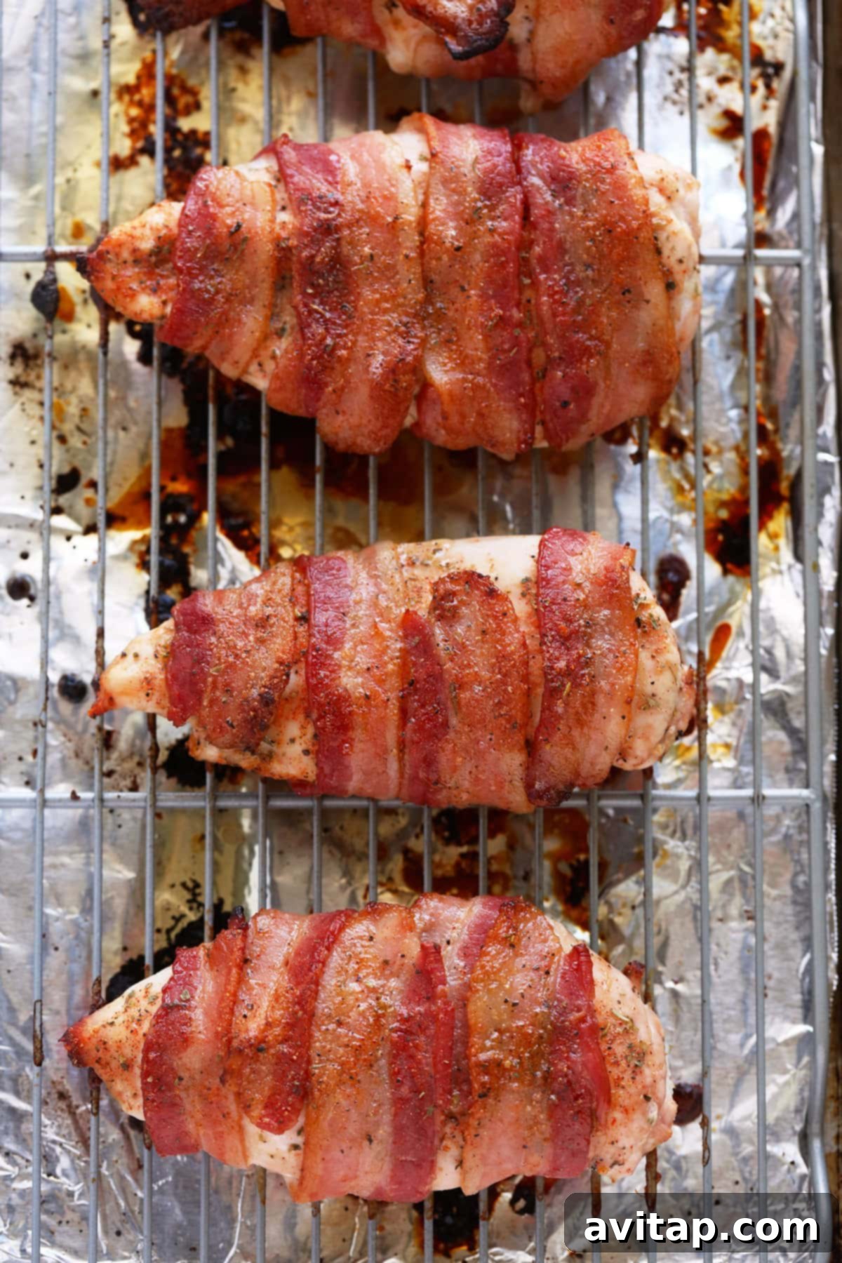 Chicken on baking rack Crispy bacon wrapped chicken breasts cooking on a metal wire rack elevated above a foil-lined baking sheet, showing the rendered grease below.