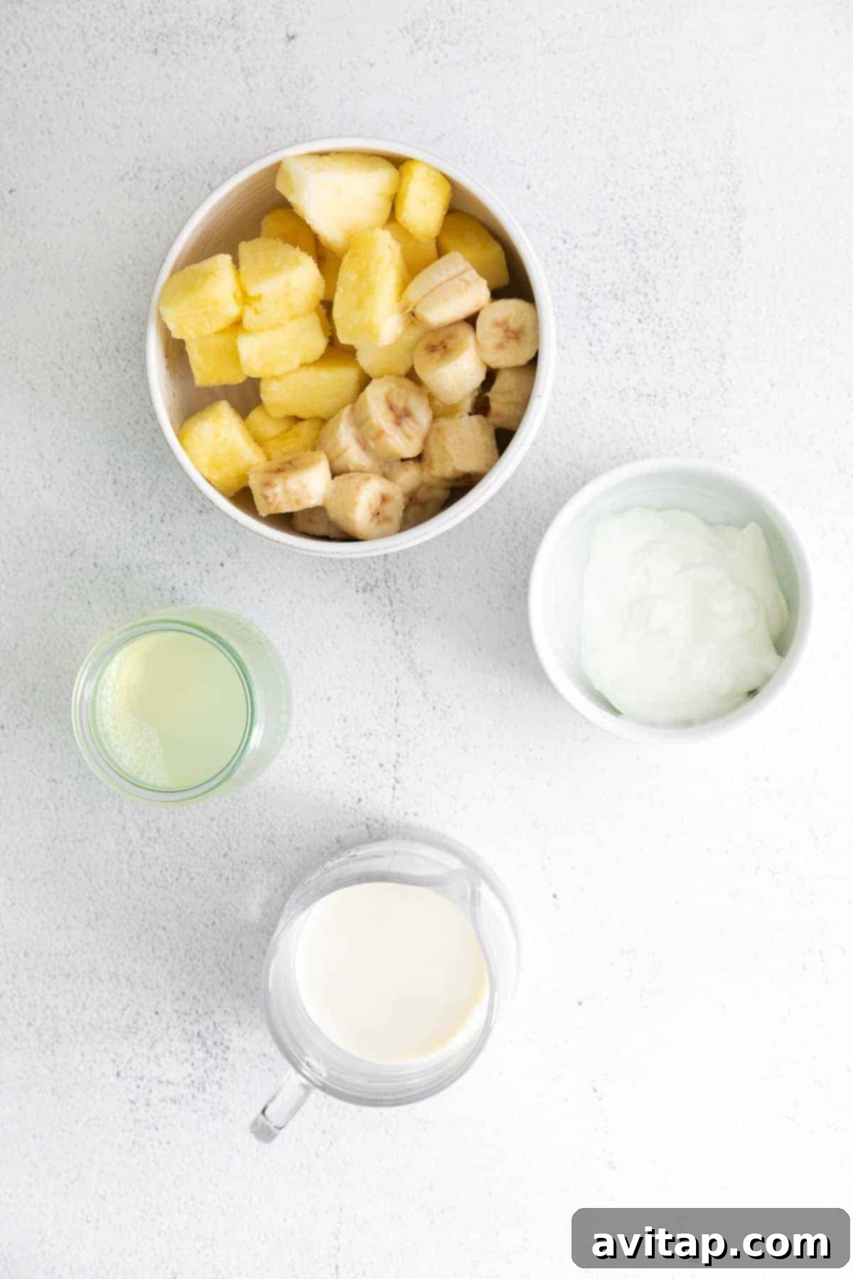 Fresh ingredients for a pineapple smoothie: frozen pineapple, banana, Greek yogurt, pineapple juice, and almond milk laid out on a kitchen countertop.