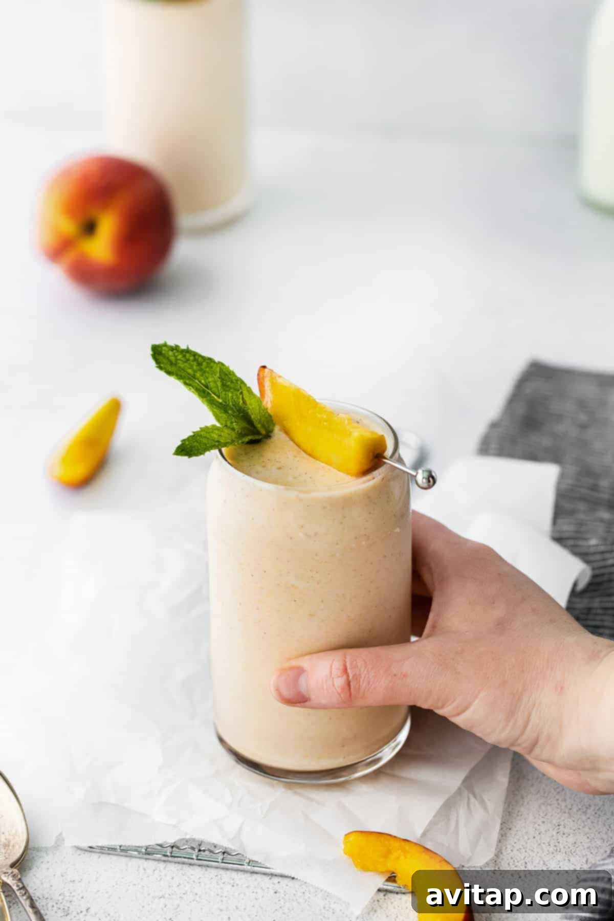 This peach smoothie tastes just like dessert, but it's healthy and made with just 5 ingredients. Make this smoothie all summer long with frozen peaches, banana, cinnamon, almond milk, and vanilla. A hand holding a tall glass of peach smoothie, garnished with a fresh peach slice, against a bright background