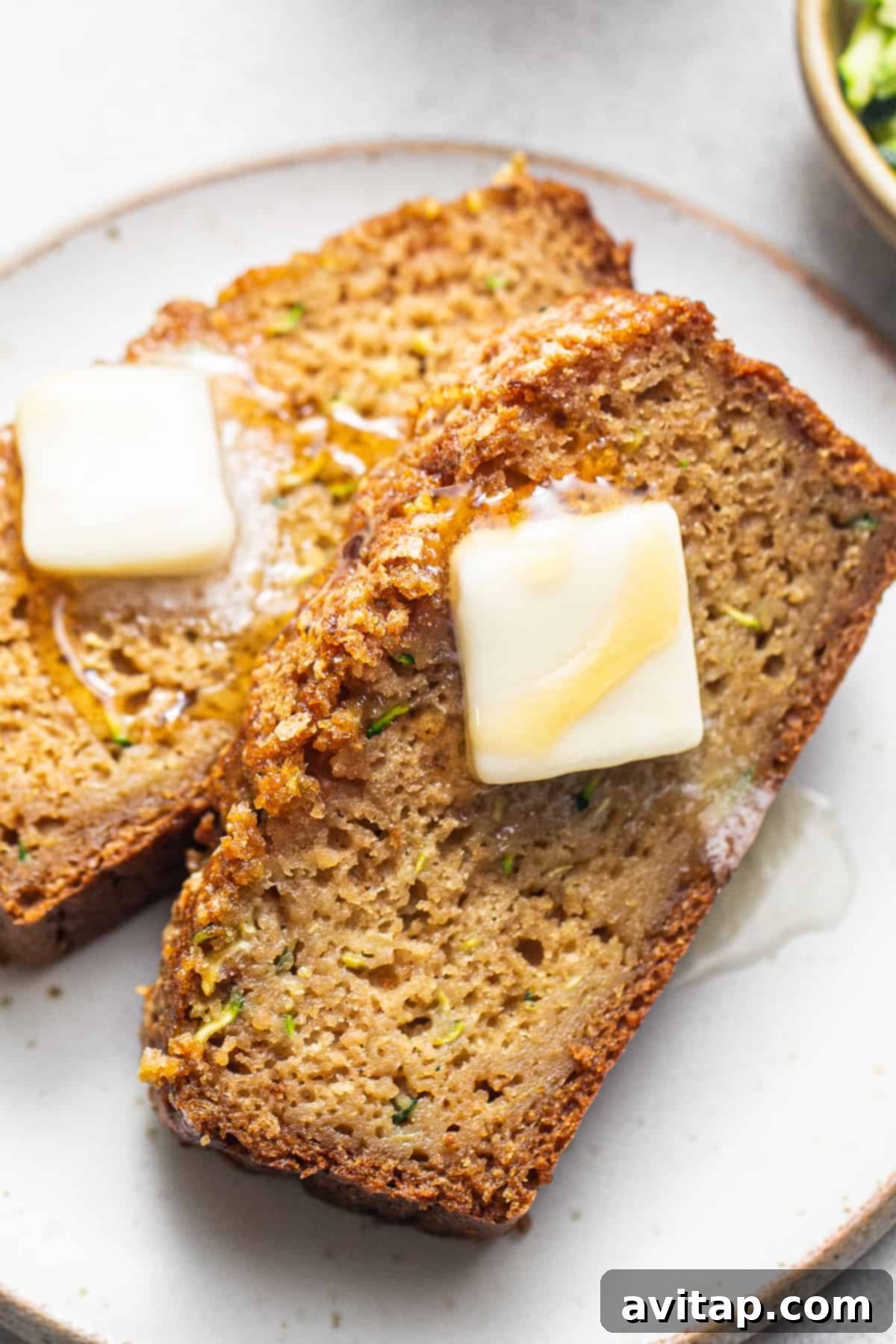 Two golden-brown slices of zucchini bread, lightly buttered, resting on a white plate, ready to be enjoyed.