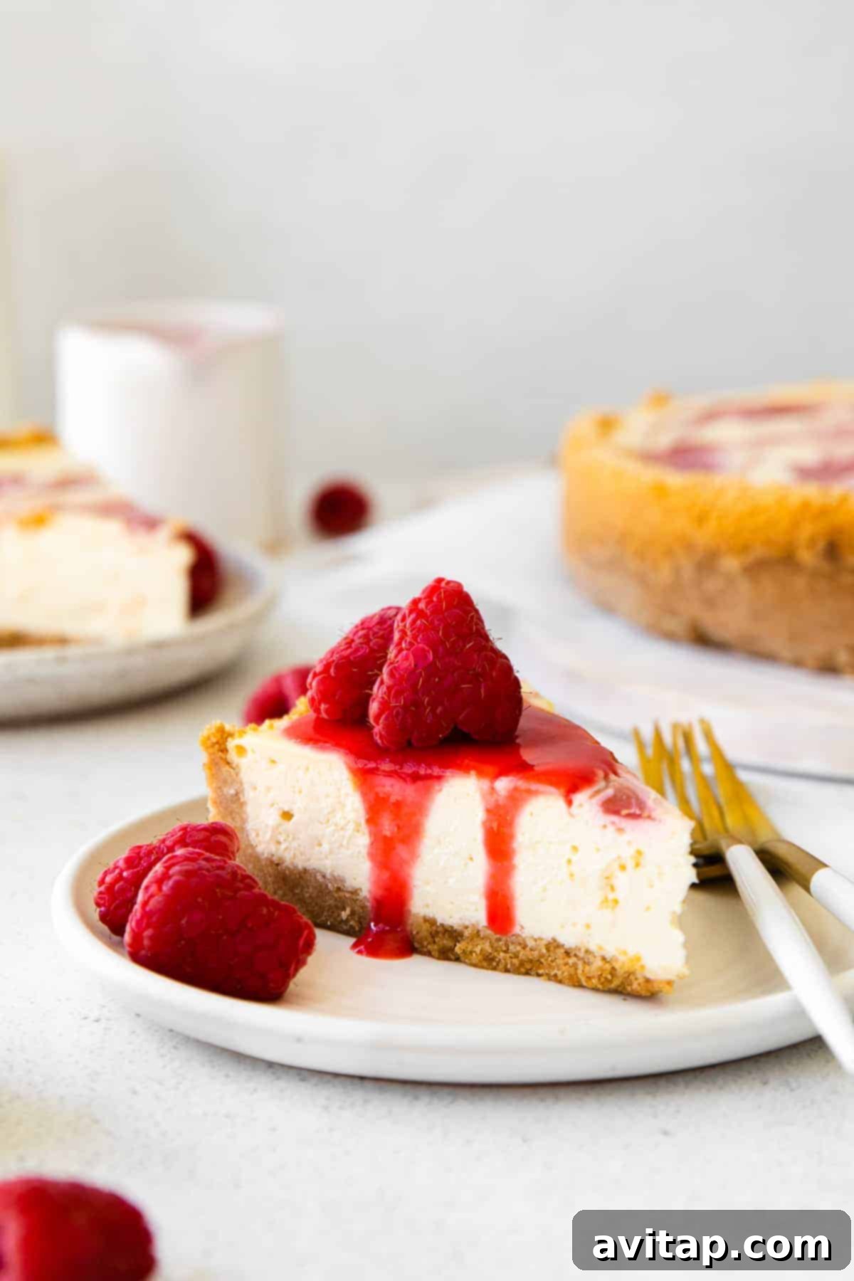 A beautiful slice of healthy cheesecake, elegantly adorned with a vibrant raspberry swirl and additional fresh raspberries, presented against a pristine white background.