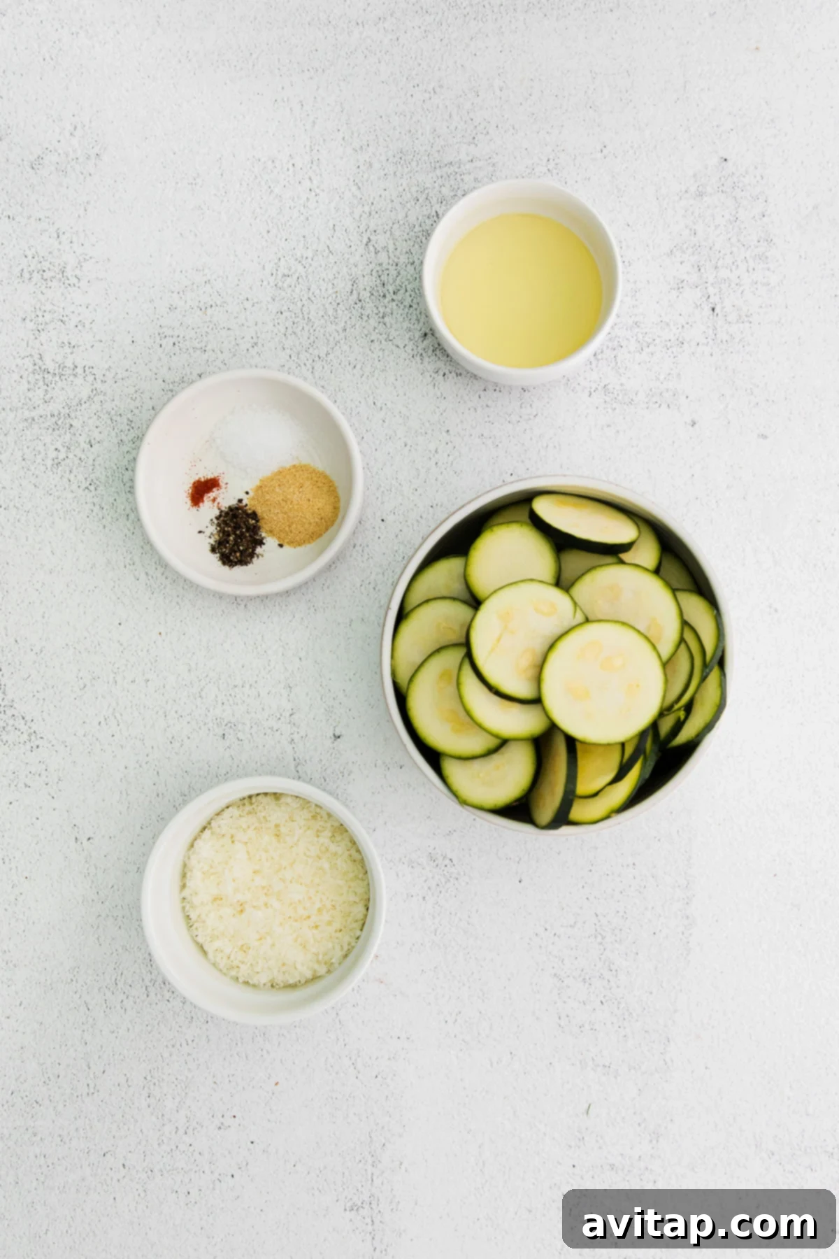 Golden Parmesan Zucchini 3 Individual bowls of olive oil, finely grated parmesan cheese, sliced zucchini, and a spice blend of garlic, salt, pepper, and cayenne pepper, ready for the baked zucchini preparation.
