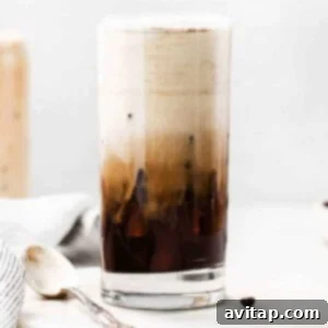 A tall glass of iced coffee on a table, topped with creamy cold foam.