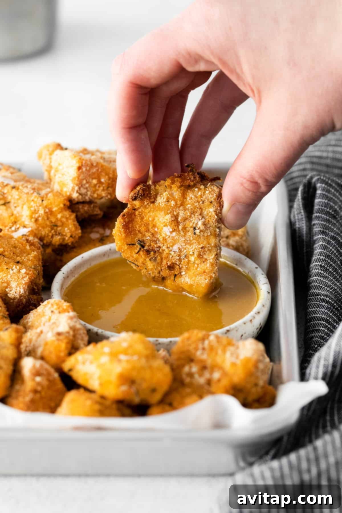 A hand dipping a crispy chicken nugget into a bowl of honey mustard sauce.
