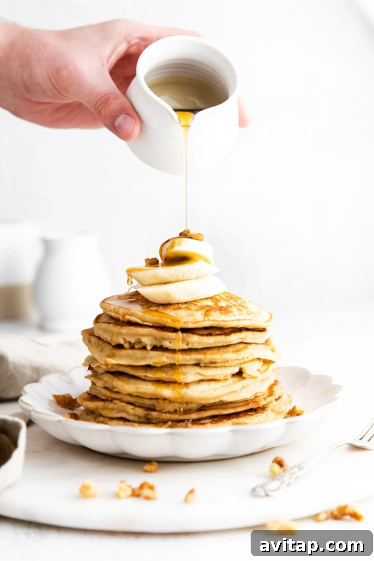 Healthy Whole Grain Banana Pancakes with Syrup Pouring maple syrup onto a stack of healthy whole grain banana pancakes with fresh banana slices on top