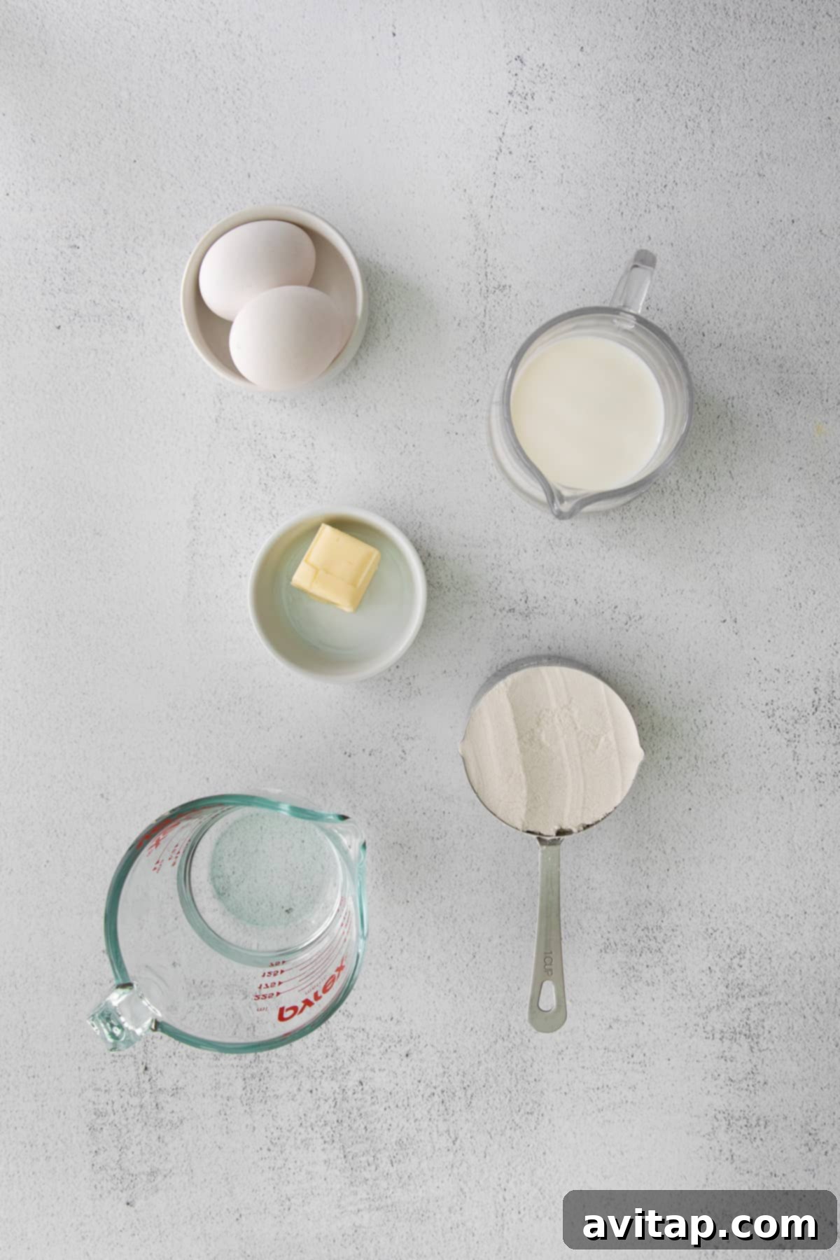 A beautiful flat lay photograph showing small white dishes filled with the raw ingredients needed for crepes: whole milk, all-purpose flour, melted butter, eggs, and water.